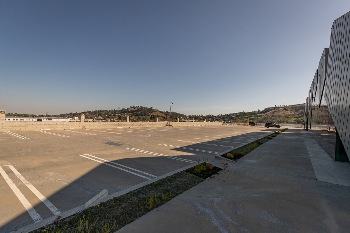 Empty rooftop parking lot with a few parked cars, shadows from a building on the right, and distant hills under a clear blue sky.