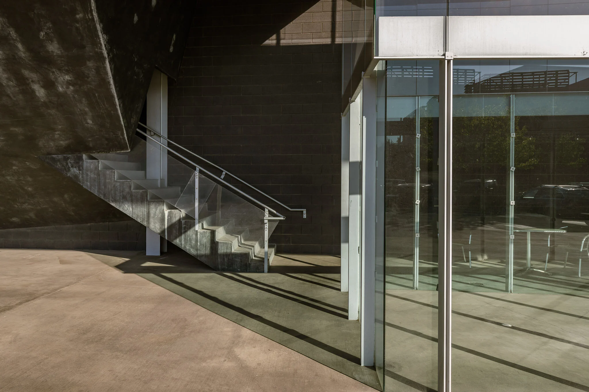 Modern building with concrete stairs, black brick wall, large glass window reflecting outside, and metal railings with shadows on the ground