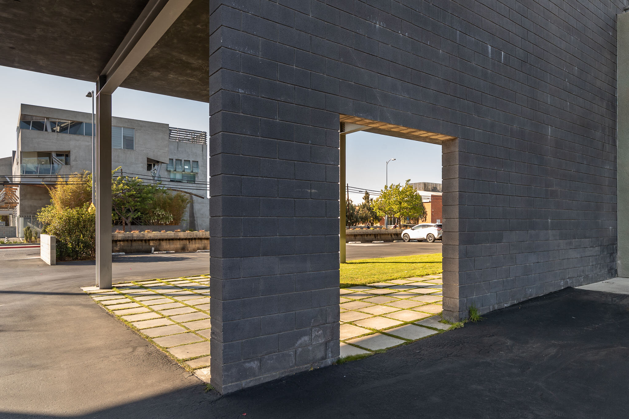 View through a modern building's rectangular opening, showing a street with a white car, trees, and a building in the background during daytime.
