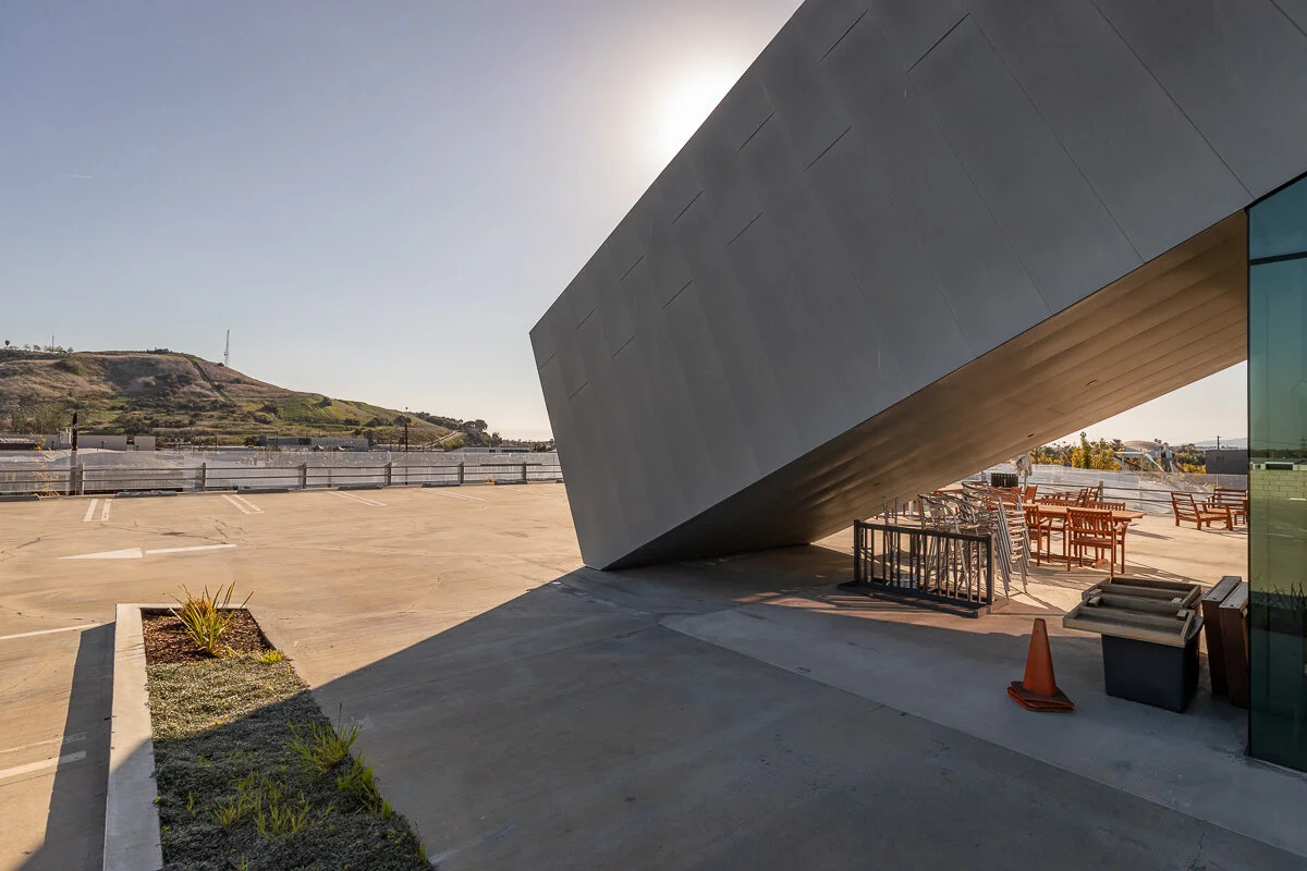 Empty outdoor parking lot with a modern building featuring a large, angled gray wall, and a row of stacked chairs and tables under the wall, with a hill and clear sky in the background.