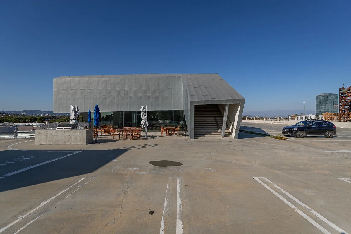 Rooftop parking lot with a modern building, outdoor seating with tables and umbrellas, a parked car, and an urban cityscape in the distance under a clear blue sky.