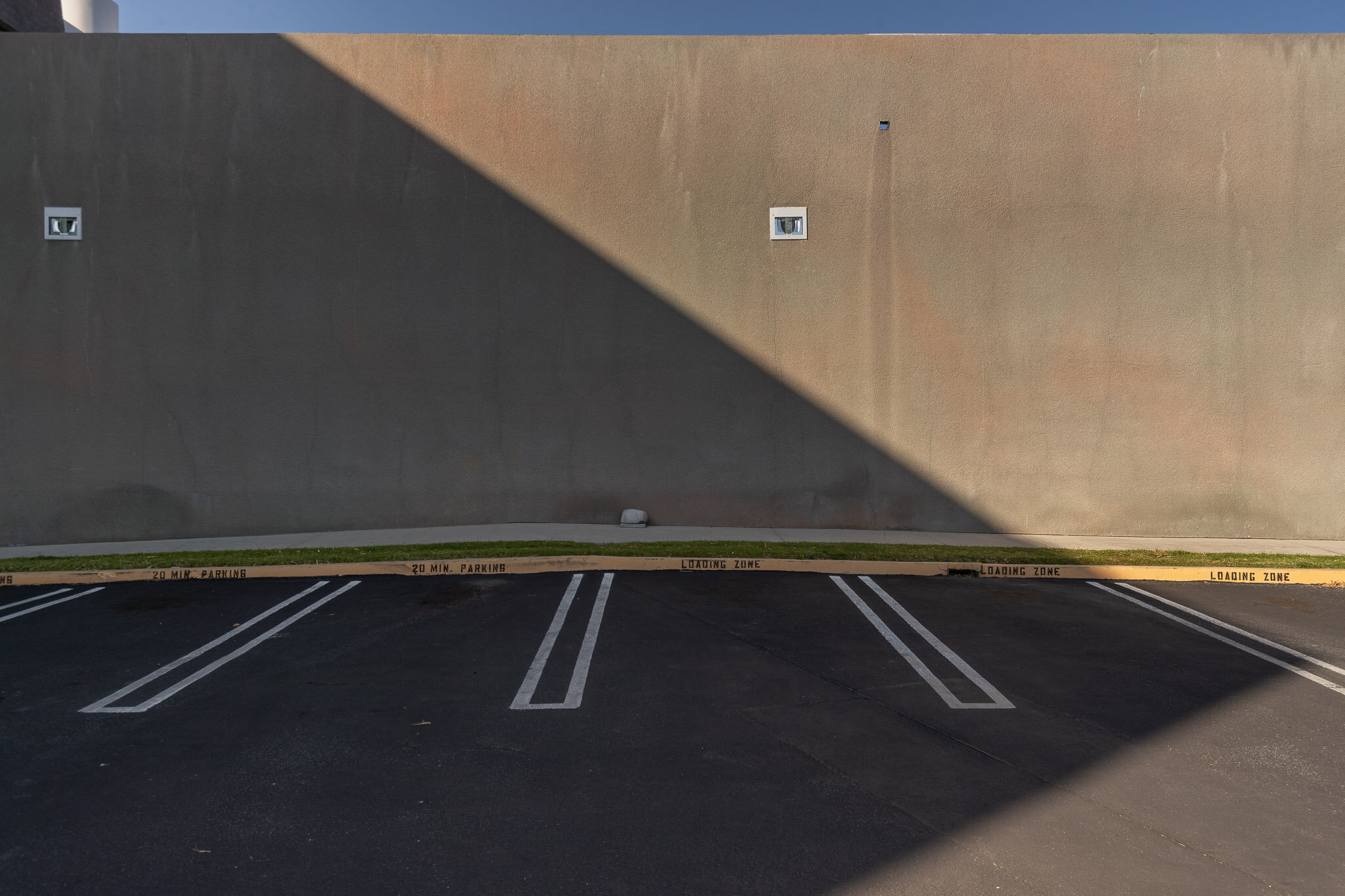 Empty parking lot with marked parking spaces, a concrete wall with small windows, and shadows cast across the ground.