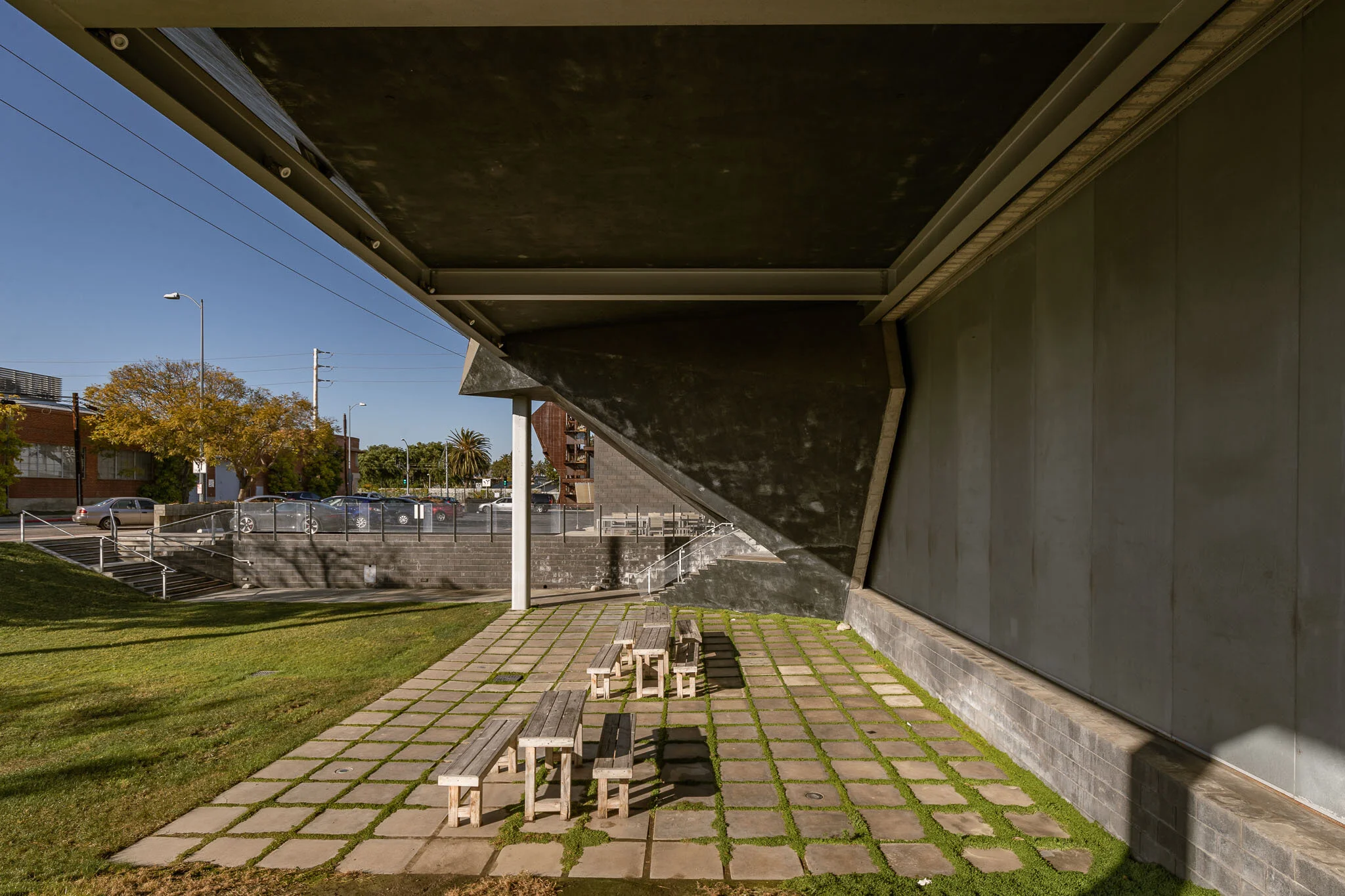 Smaller wooden benches and tables on concrete pavers outside under a large building overhang, with cars and trees in the background.