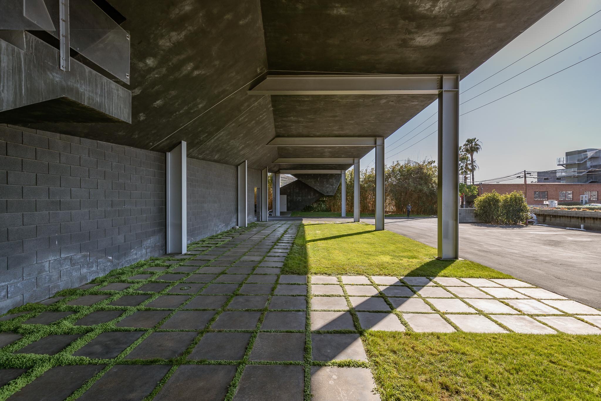 Modern building exterior with covered walkway, concrete slabs, grass patches, and support columns under a concrete overhang.