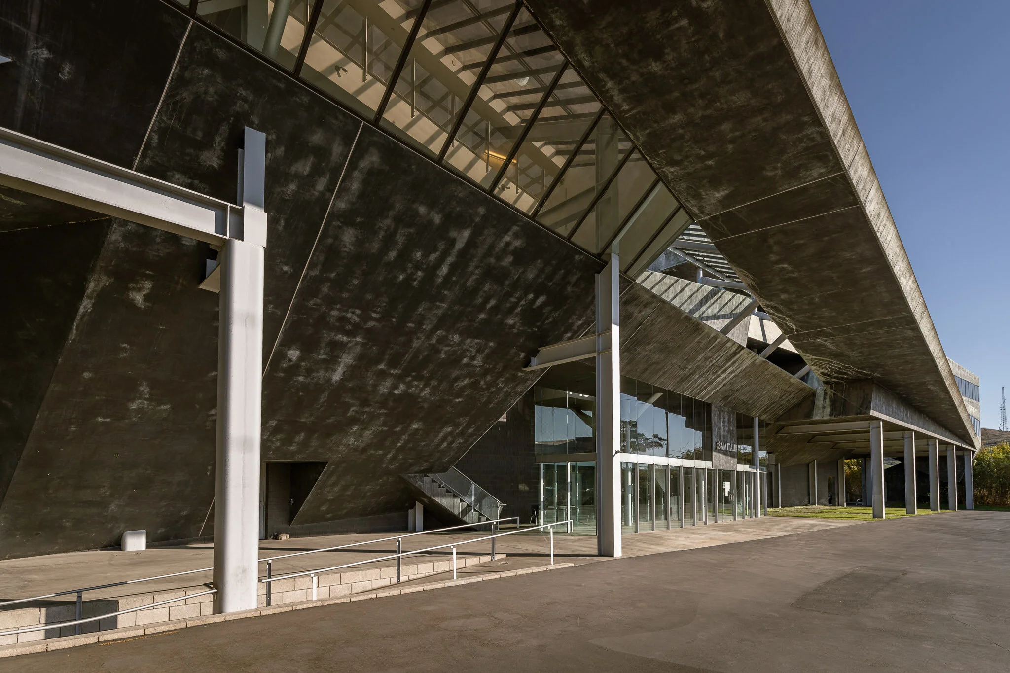 Modern building with dark concrete exterior, glass windows, and an overhanging upper floor, with a concrete sidewalk and stairs leading to the entrance.