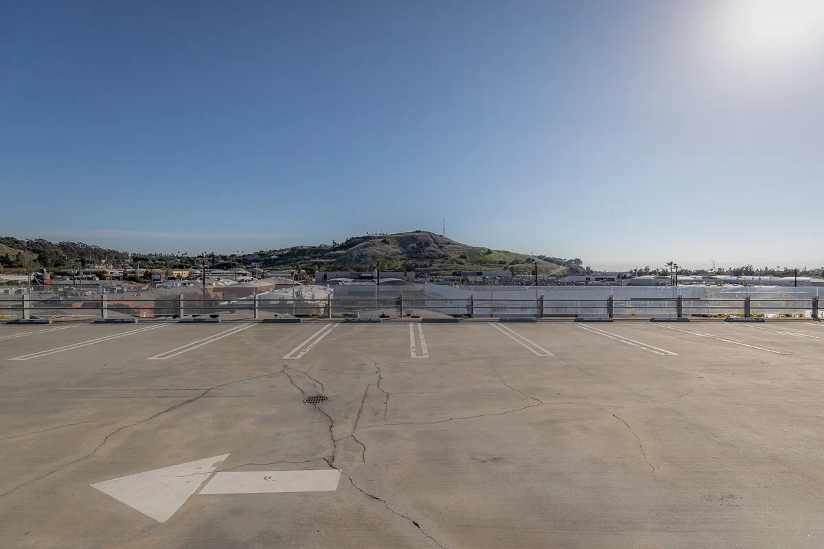 Empty rooftop parking lot with a white arrow painted on the concrete pointing left, overlooking a harbor with boats and a hilly landscape in the background under a clear sky.