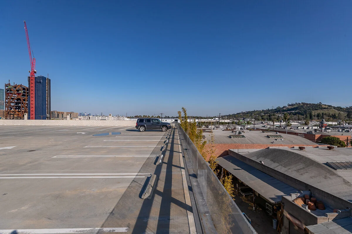 Empty rooftop parking lot with one parked vehicle, under a clear blue sky, adjacent to a cityscape with tall buildings and a hill.