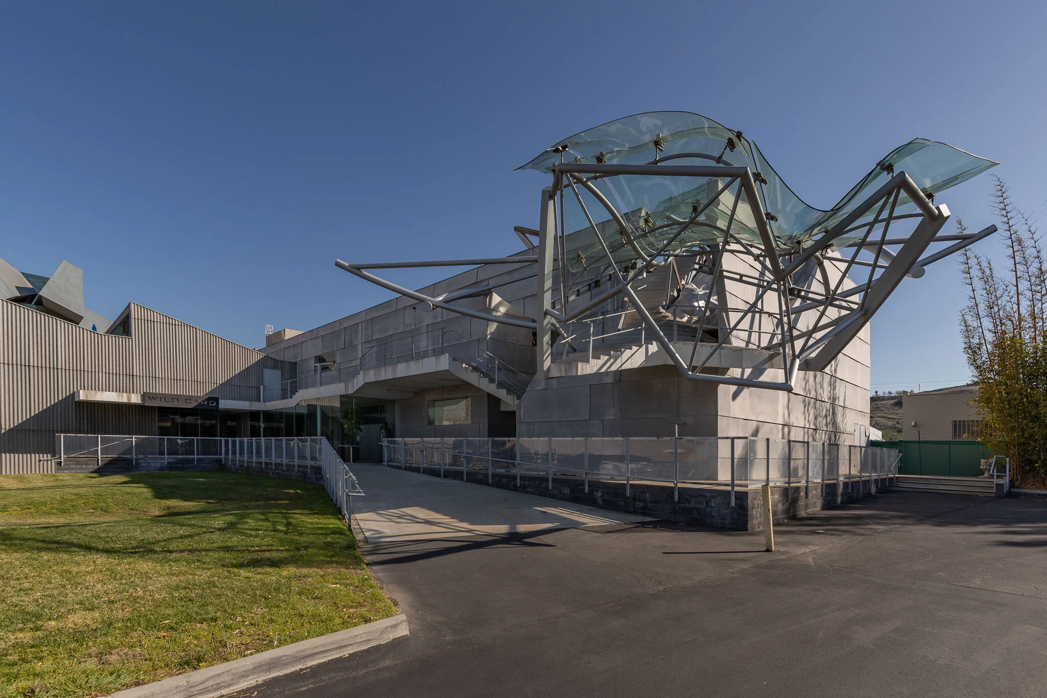 Modern building with unique glass and metal sculpture on roof, concrete and glass walls, and a ramp leading to the entrance, under a clear blue sky.