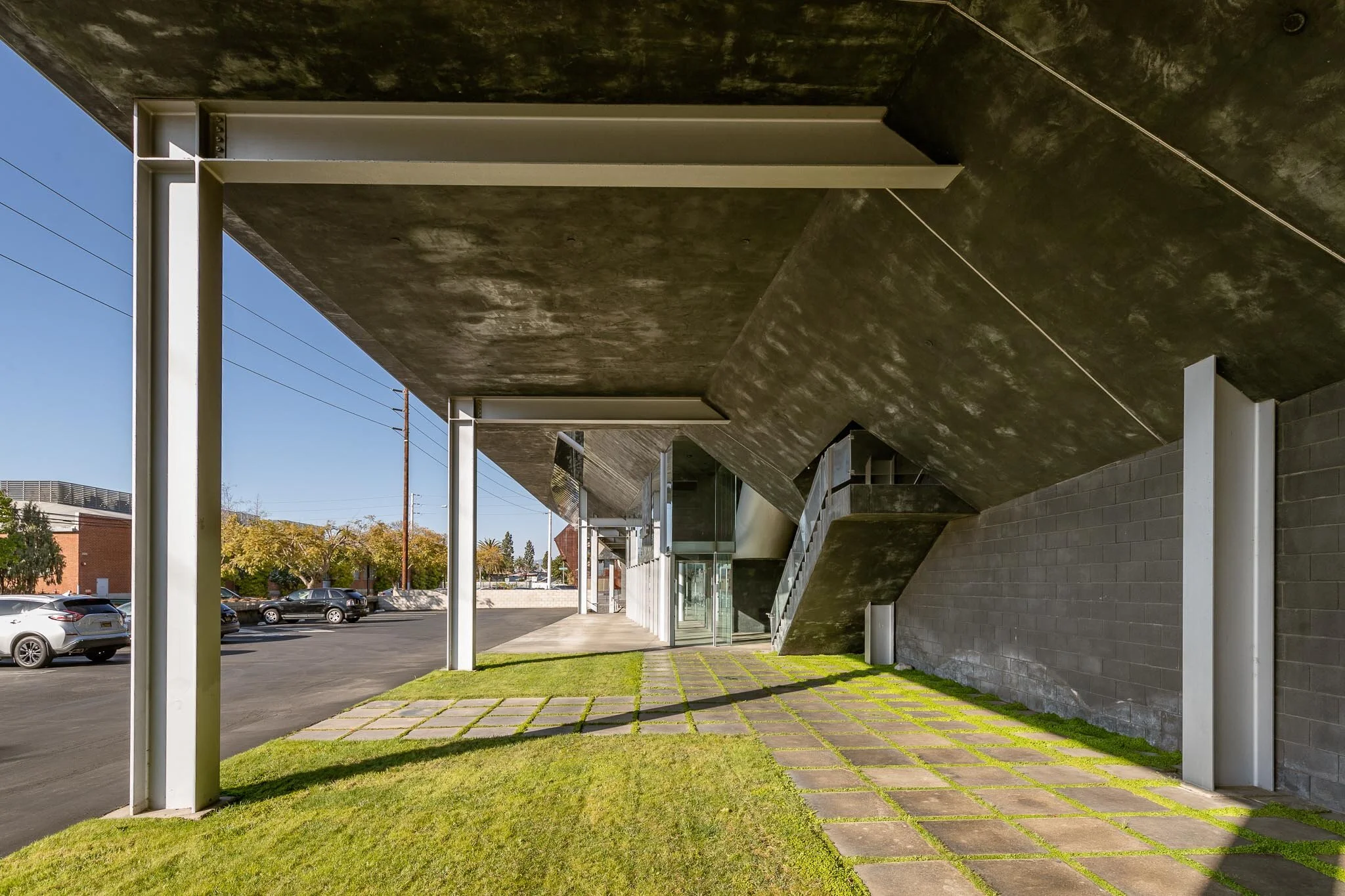 Modern building exterior with overhang, grassy walkway, and parking lot in the background.