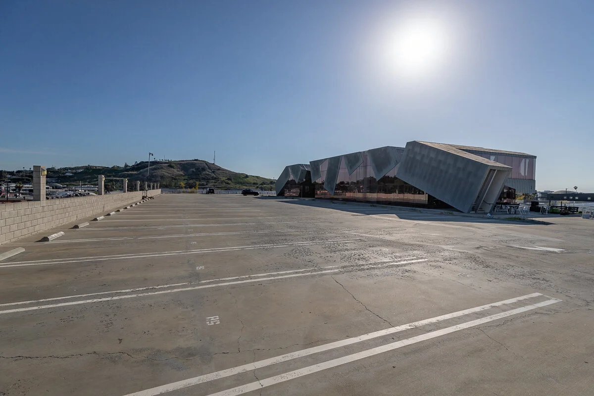 Empty parking lot with a modern building featuring angular, reflective glass panels in the background and a hill with scattered vegetation in the distance.
