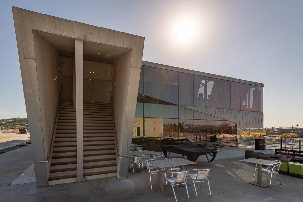 Modern building with glass exterior and an exterior staircase leading to an upper level, outdoor seating area with tables and chairs, under a bright sky with the sun shining.