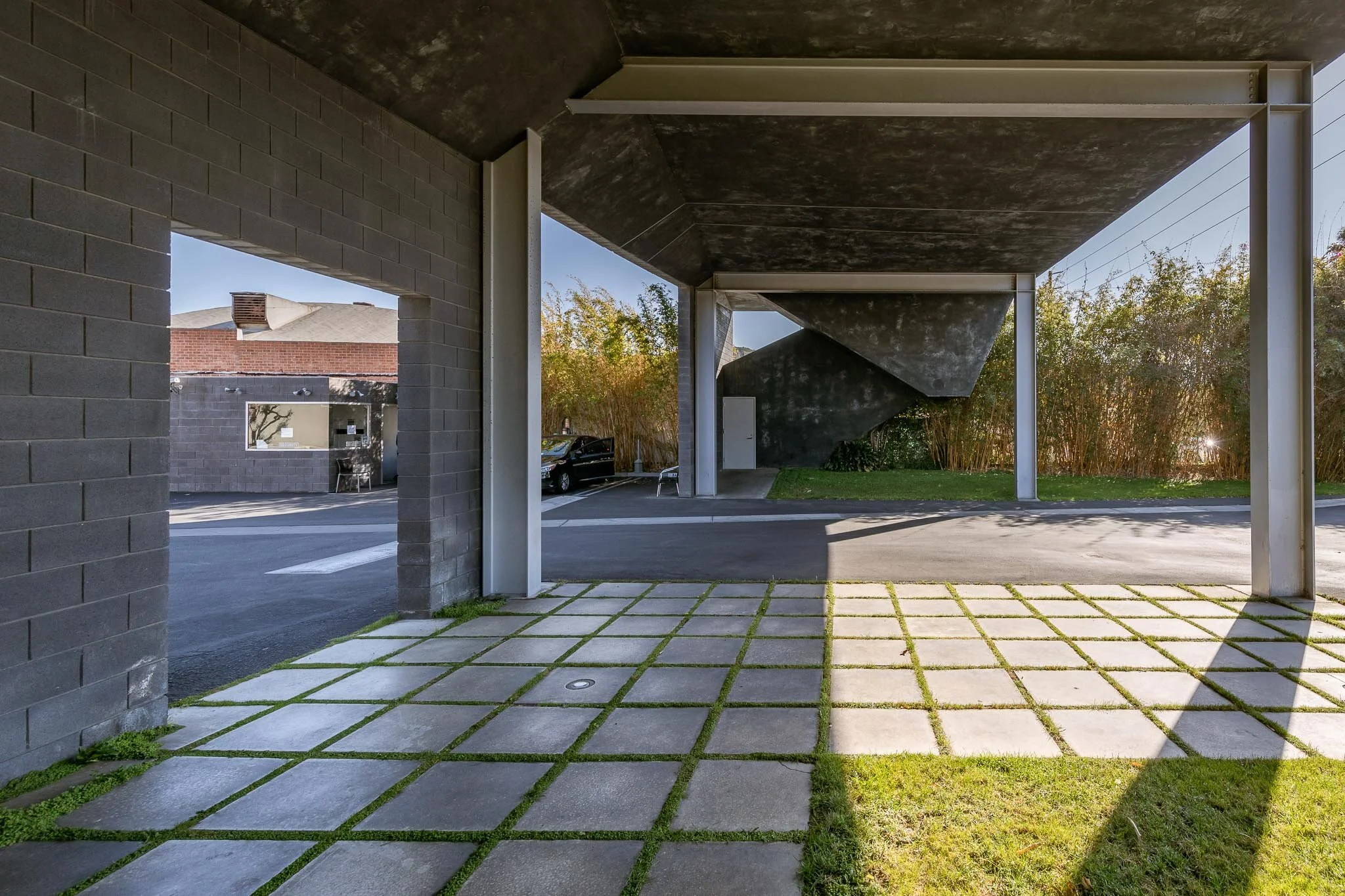 Modern building exterior with gray brick walls, concrete pavement with grass in between, shadow cast on ground, parked car, staircase, and foliage in background