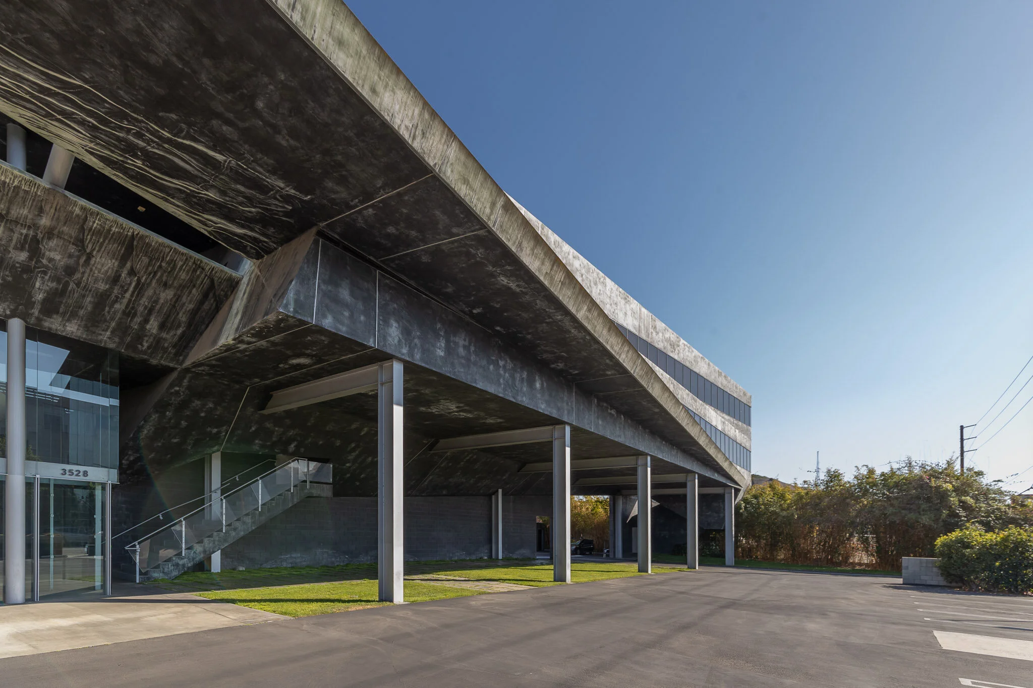 Modern commercial building with an overhanging concrete structure, metal support pillars, glass windows, stairs, and a parking lot, under a clear blue sky.