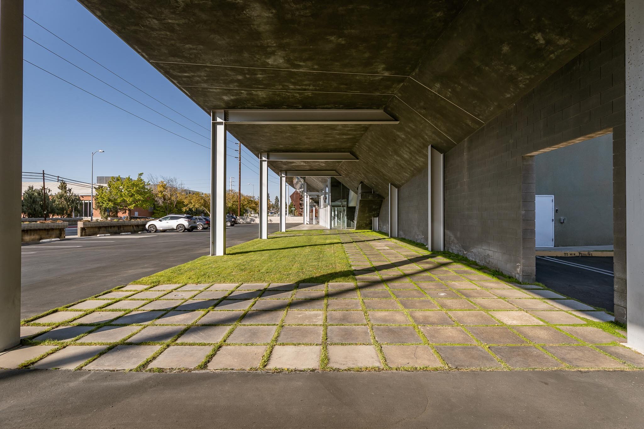 Sidewalk with interlocking concrete pavers and grass patches underneath an overhanging building structure, with parked cars and trees in the background