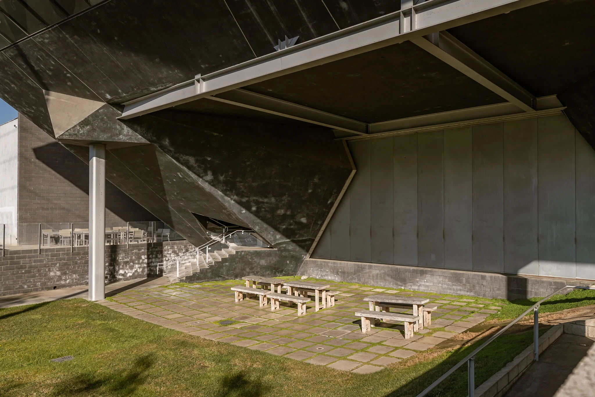 Outdoor area with concrete paving, wooden benches, and a modern, angular building structure with overhangs and stairs.