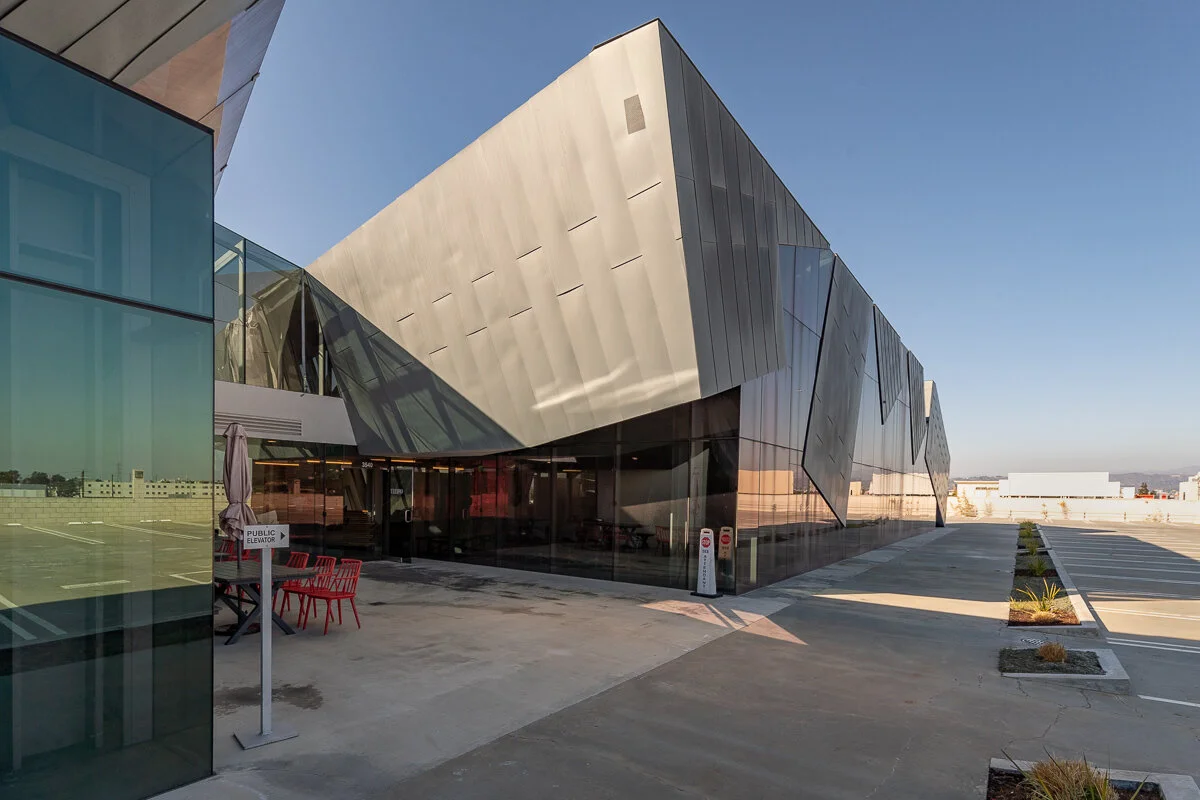 Modern architectural building with angular metallic and glass exterior, sunny day, empty parking lot in foreground.