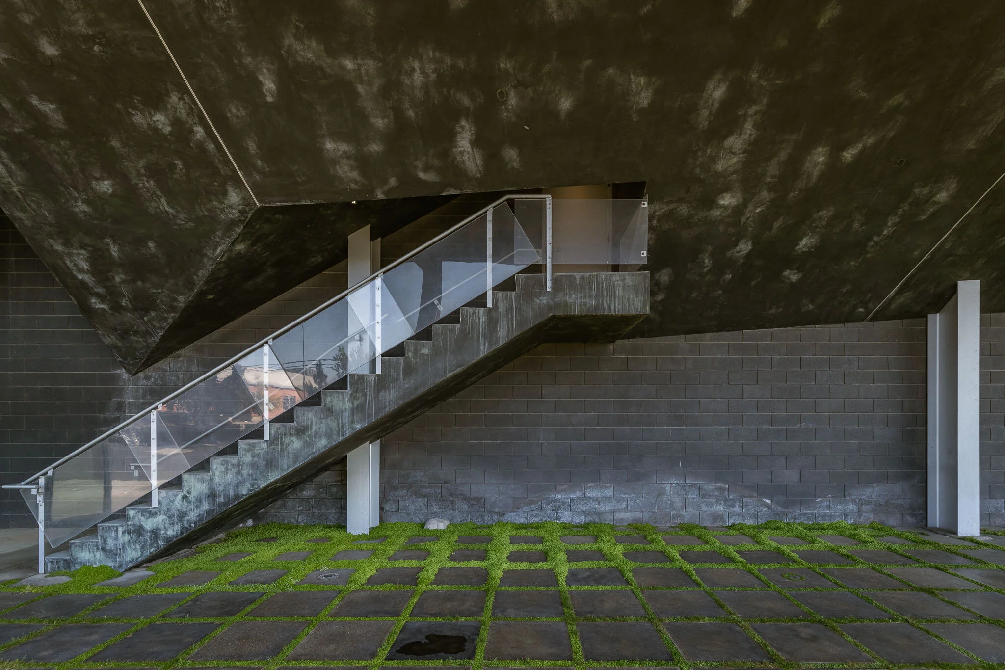 Modern outdoor staircase with glass railing against a dark brick wall, with a ground pattern of square concrete pavers separated by grass.