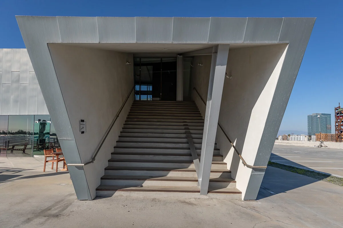 Modern, angular staircase leading to an entrance on a building rooftop with clear blue sky and cityscape in the background.