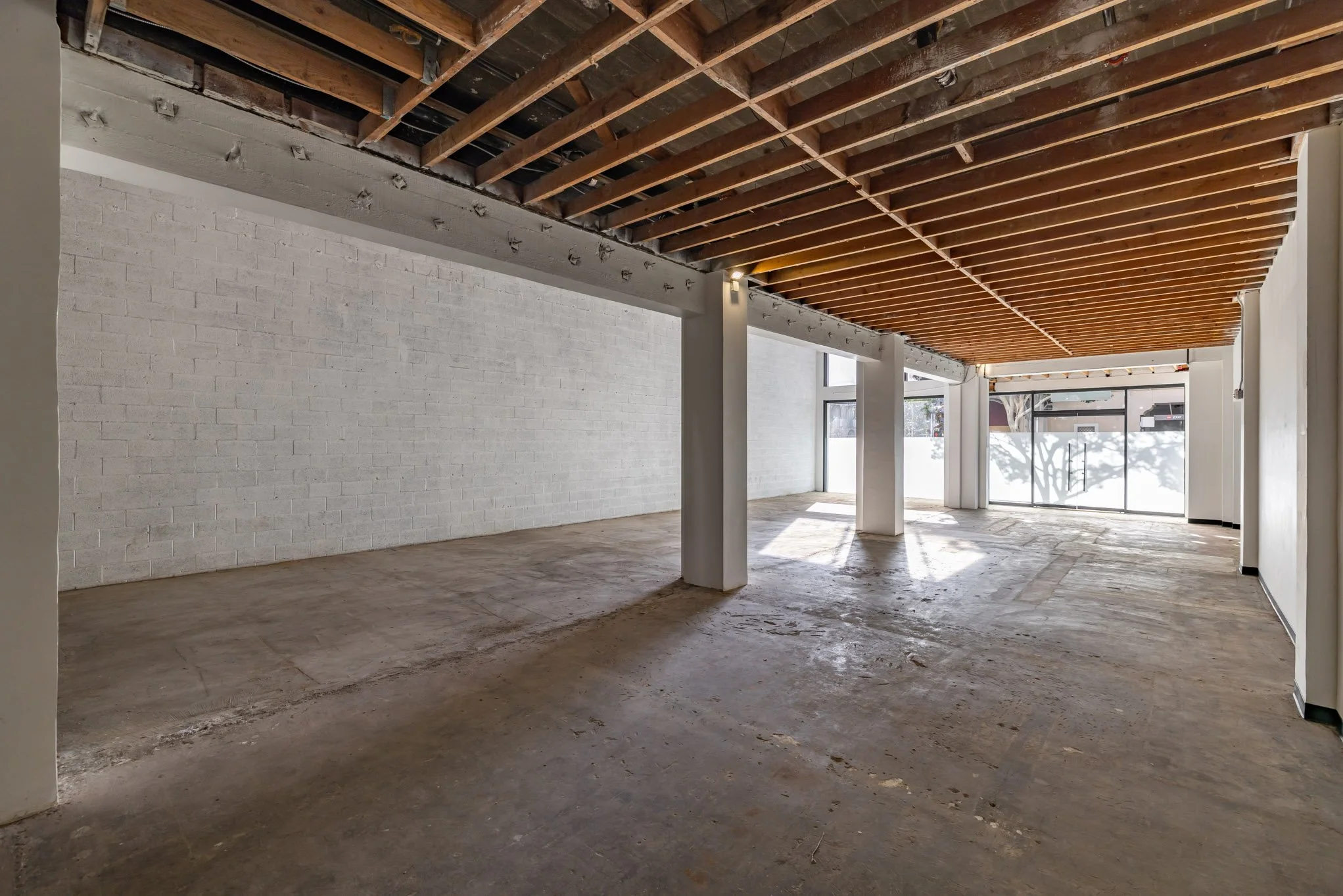 Empty commercial space with white brick walls, exposed wooden ceiling, concrete floor, and large glass storefront windows with natural light.