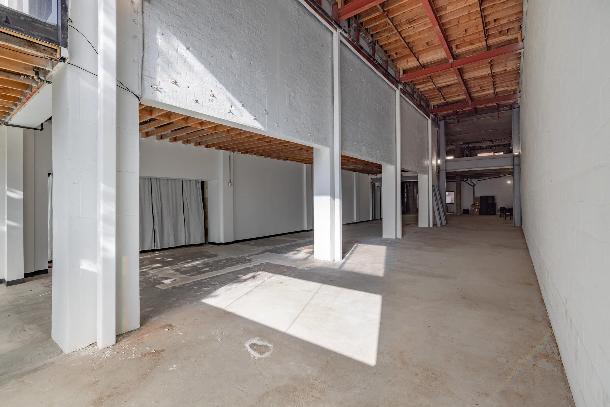 Empty commercial interior space under construction with concrete floors, white walls, exposed ceiling with wooden beams, and support columns.