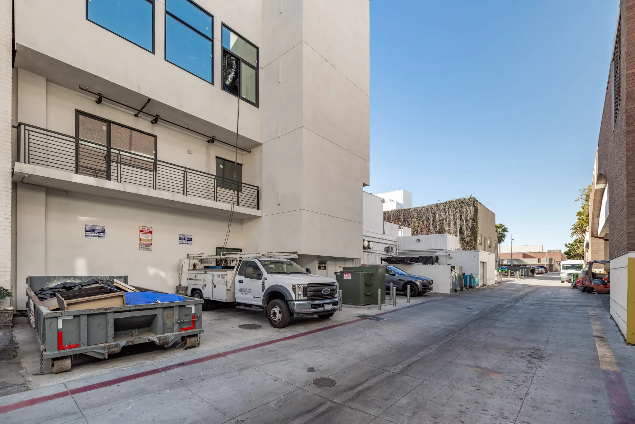An alleyway with parked commercial vehicles, including a white utility truck, and trash dumpsters next to multistory modern buildings under a clear blue sky.