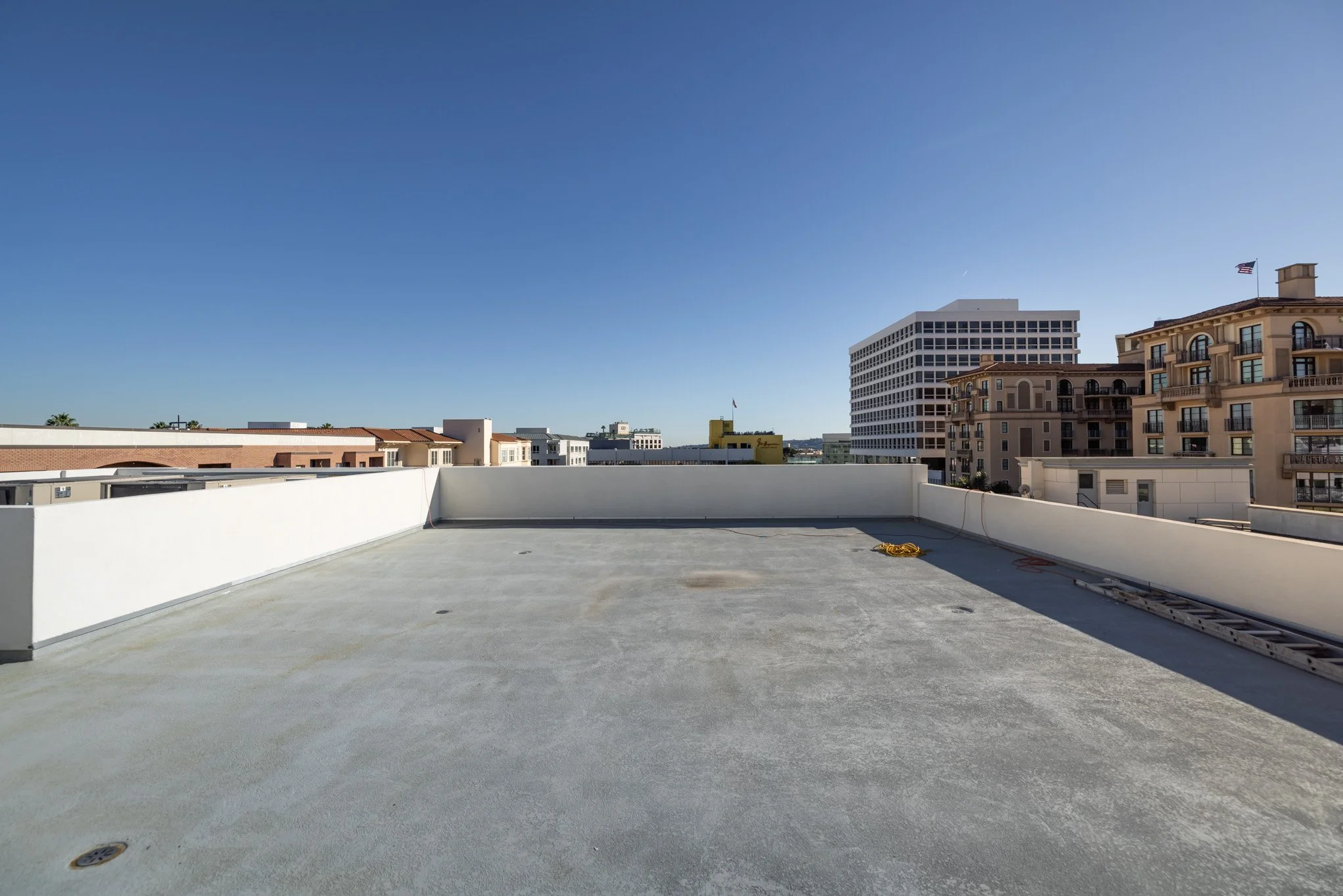 Empty rooftop with a white parapet wall, yellow construction rope, and a ladder, overlooking a city with high-rise buildings under a clear blue sky.