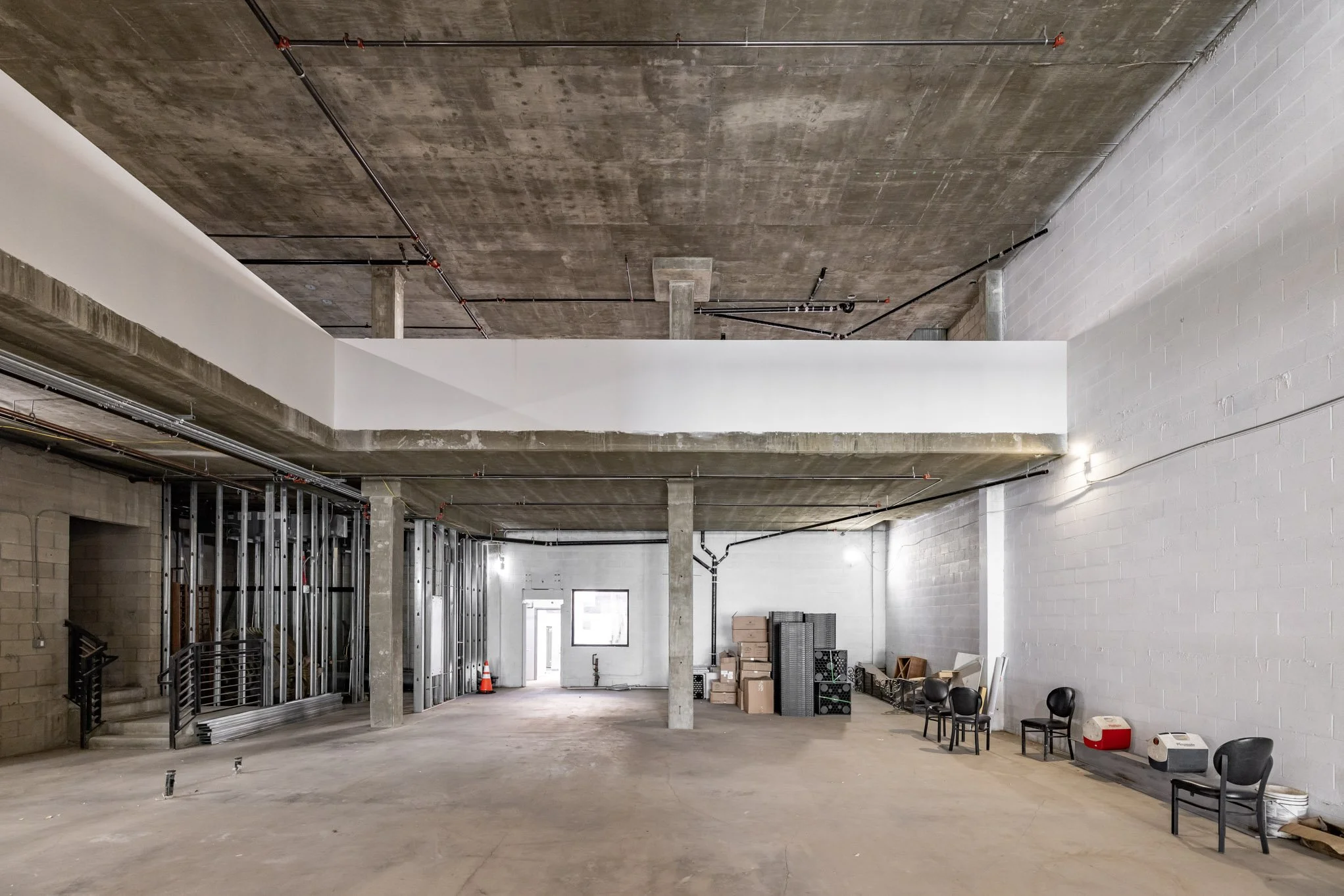 Empty interior of a building under construction or renovation, with exposed concrete floors, walls, and ceiling, with some stacked boxes and chairs along the right wall, and construction materials on the left.