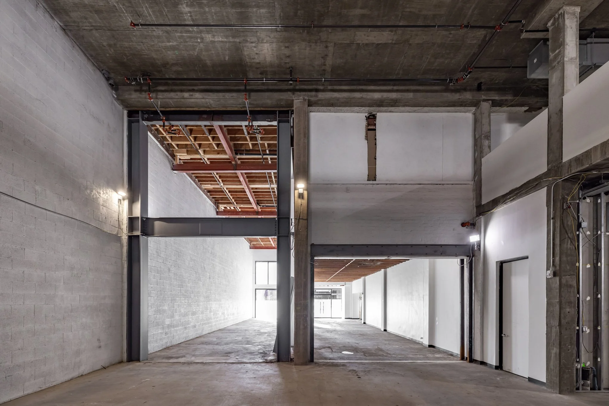 Interior view of an unfinished building with exposed concrete walls, a concrete ceiling, and construction wiring. Sections of the ceiling reveal wooden framing and ceiling structures. Natural light enters from large windows in the background.