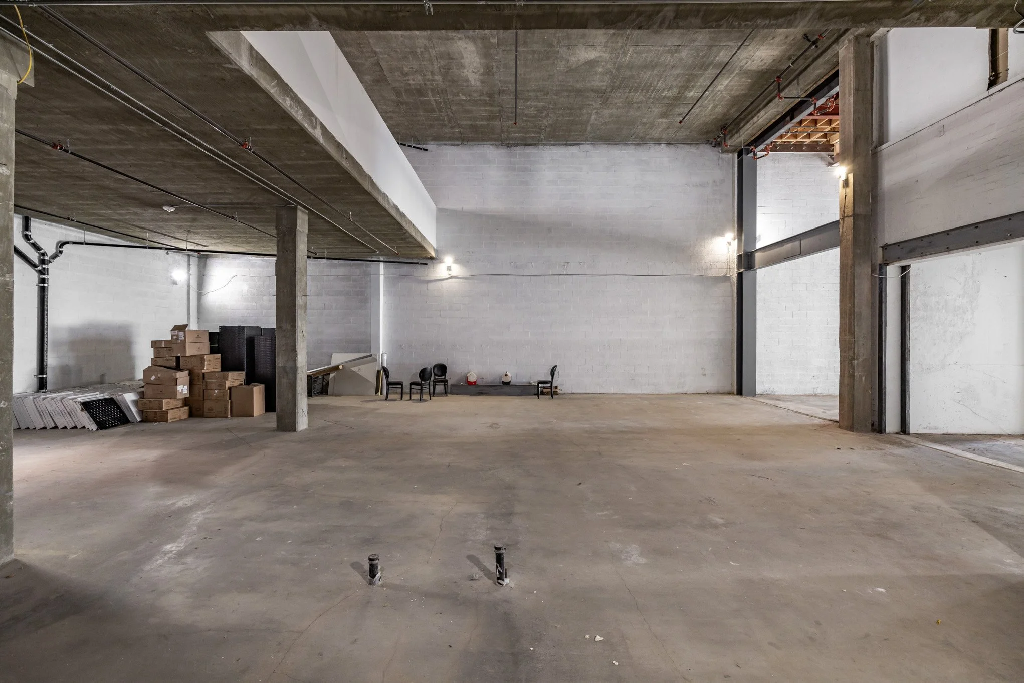 An unfinished indoor space with concrete floors, exposed concrete ceiling, some furniture and boxes against a white cinder block wall.