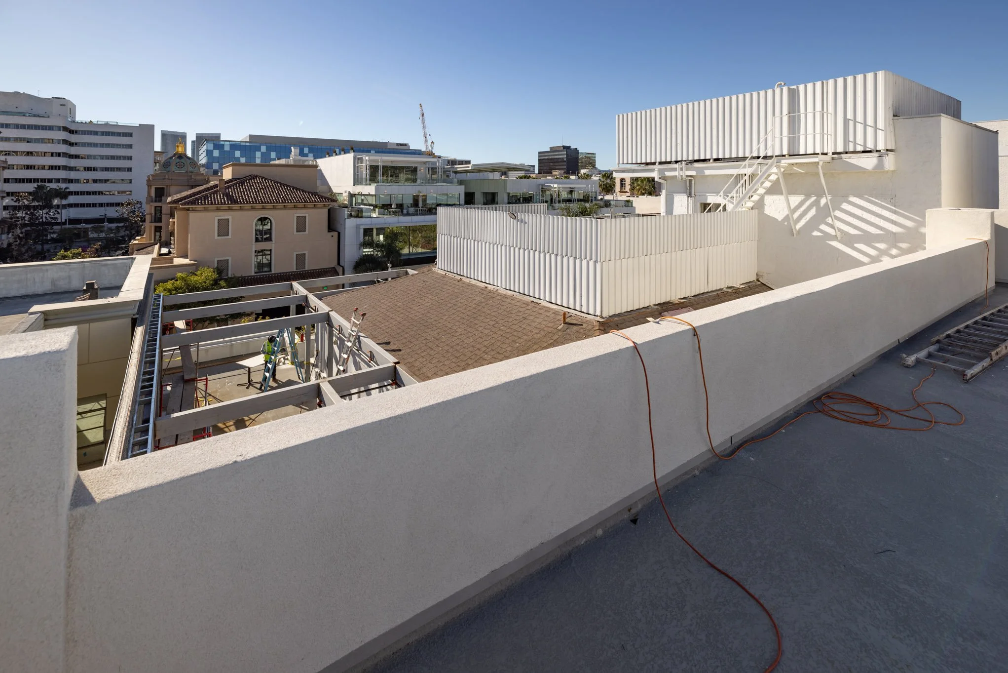 Rooftop construction scene showing ladders, orange extension cords, and a building with various neighboring structures against a clear blue sky.