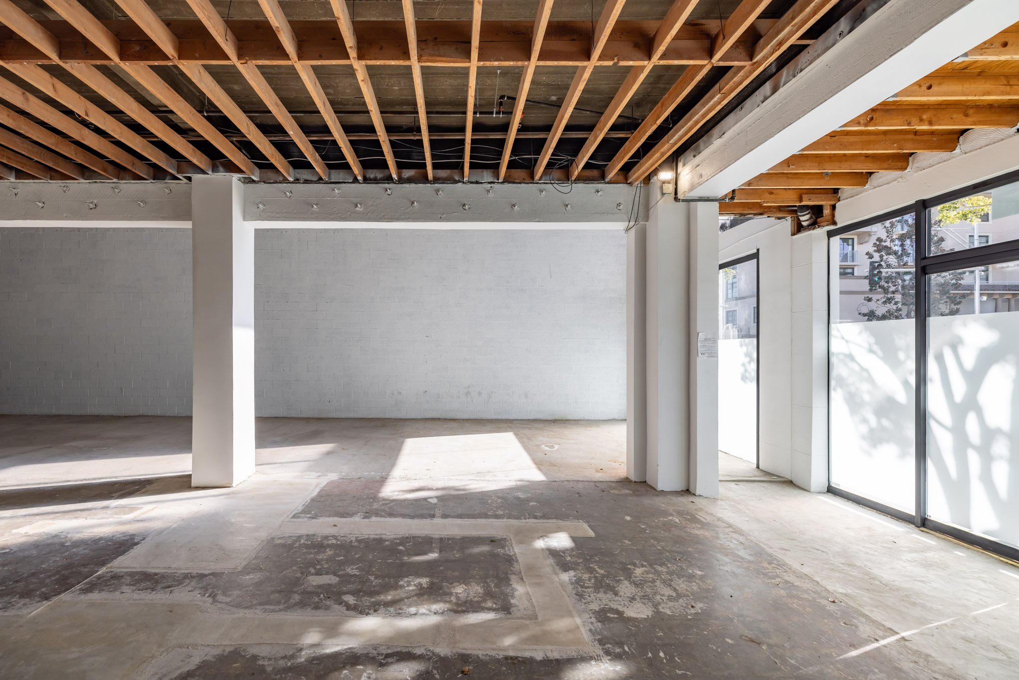Interior of an unfinished commercial space with exposed wooden ceiling beams, a white brick wall, and large glass windows and doors letting in natural light.