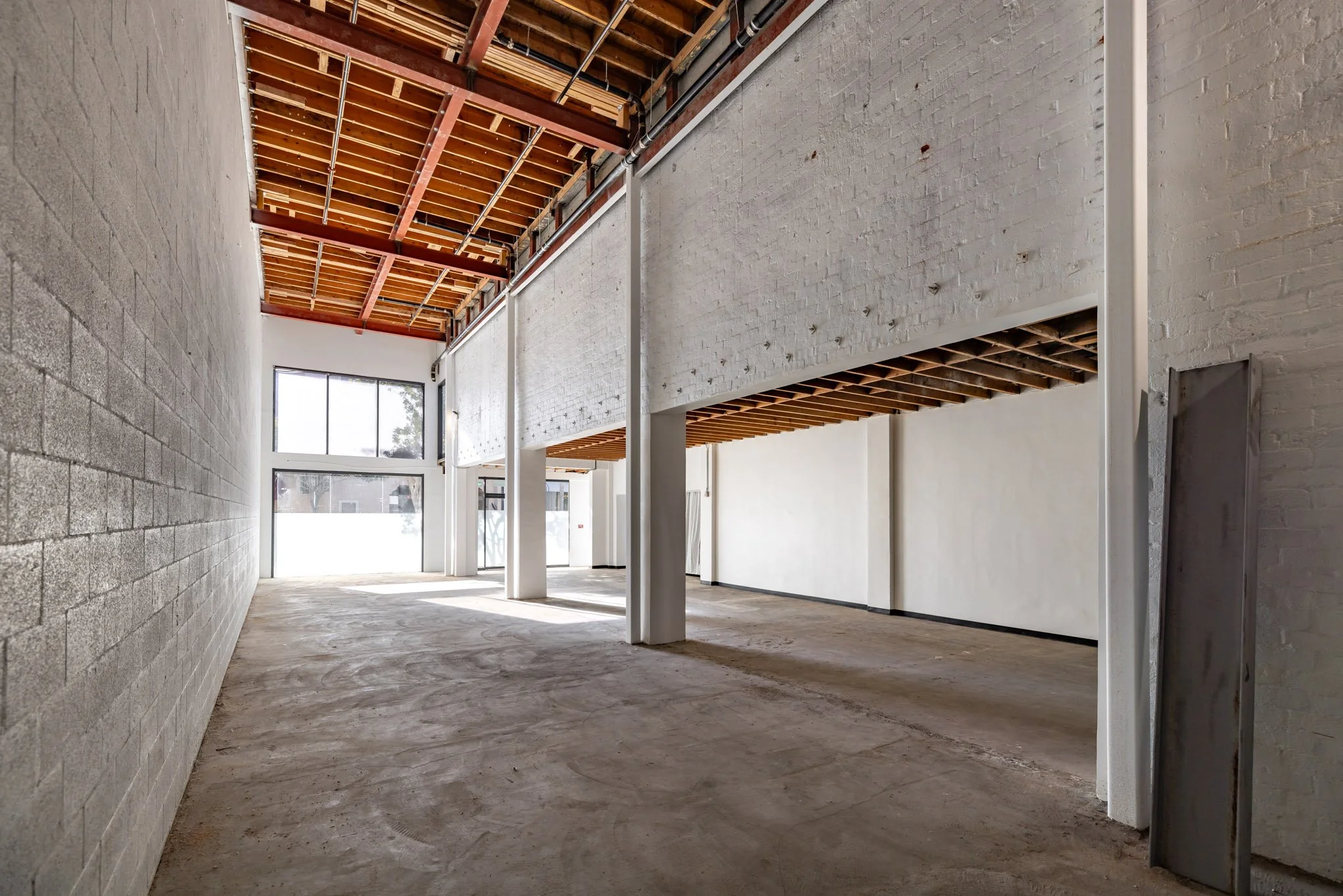 Interior view of an unfinished commercial space with concrete floors, white brick walls, large front windows, and a high ceiling with exposed wooden beams and metal supports.