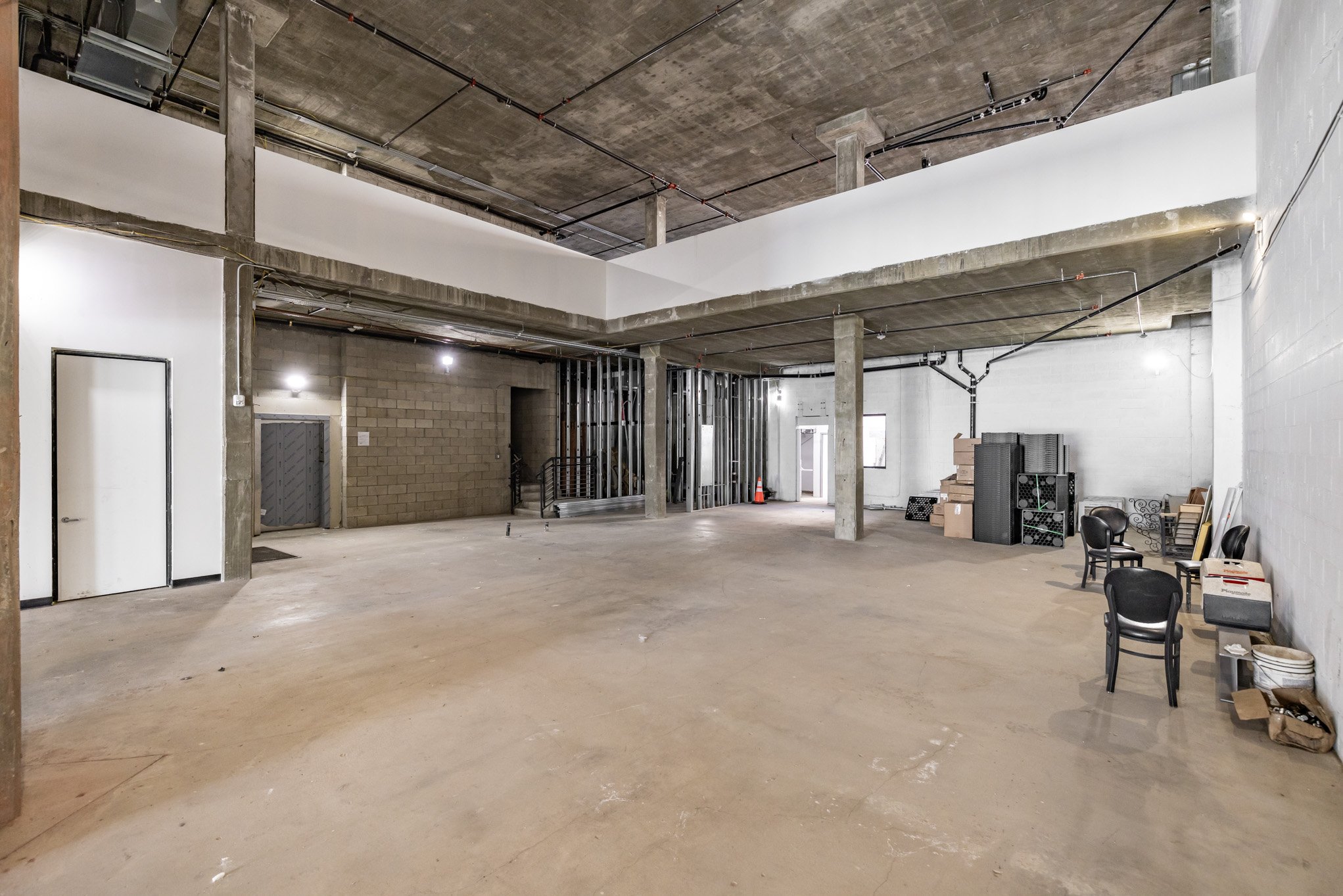 Large, unfinished interior space with concrete floors, exposed ceiling, and walls. Some chairs, boxes, and equipment are stored along the walls.