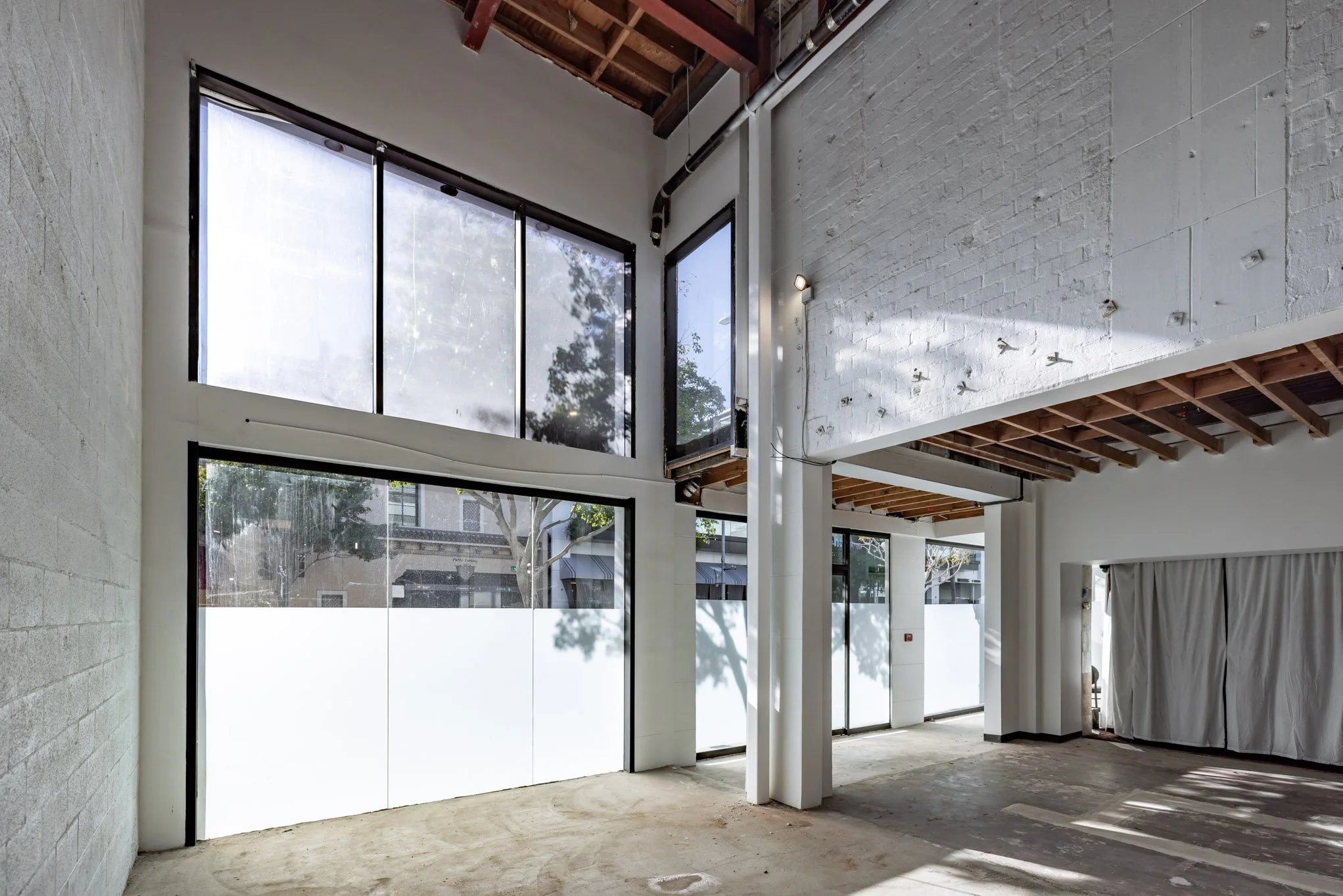Interior of an unfinished building with large windows, white walls, exposed ceiling beams, and a concrete floor.