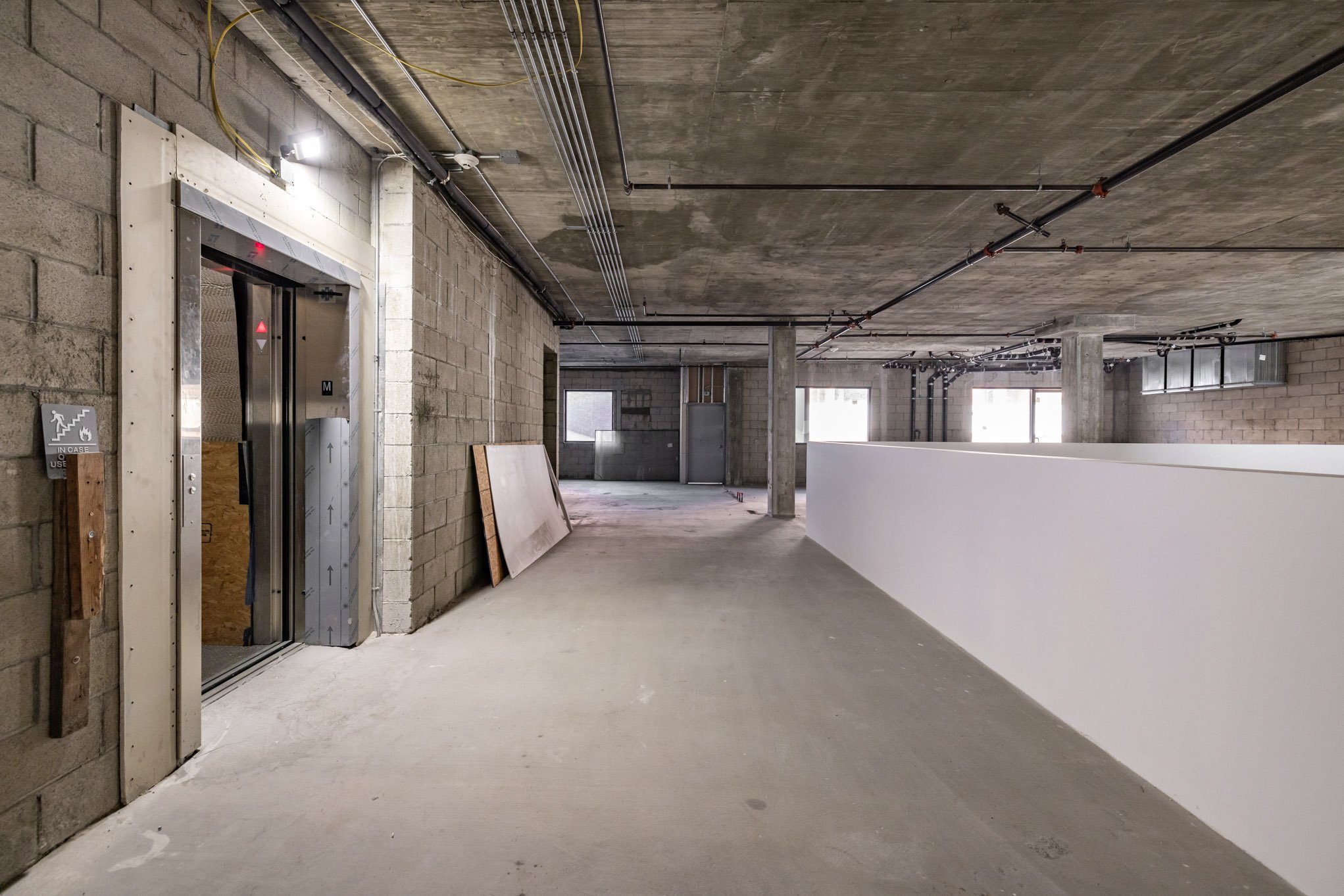 Empty interior space of a building under construction with exposed concrete ceiling, walls, and electrical wiring, including an elevator on the left side.
