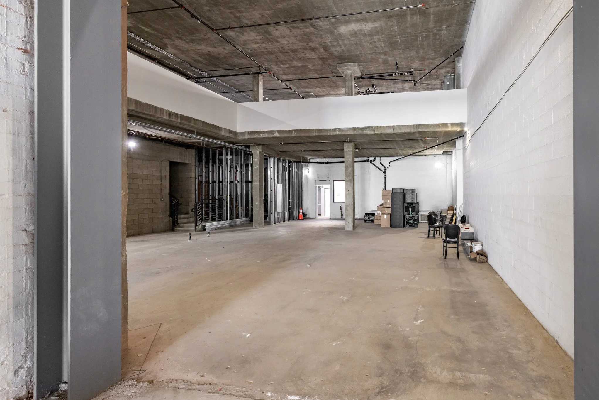 Interior of a building under construction or renovation with exposed concrete floors, white and gray walls, and ceiling. There are construction materials, chairs, and boxes along the far wall. Metal framing and ducts are visible on the upper level an