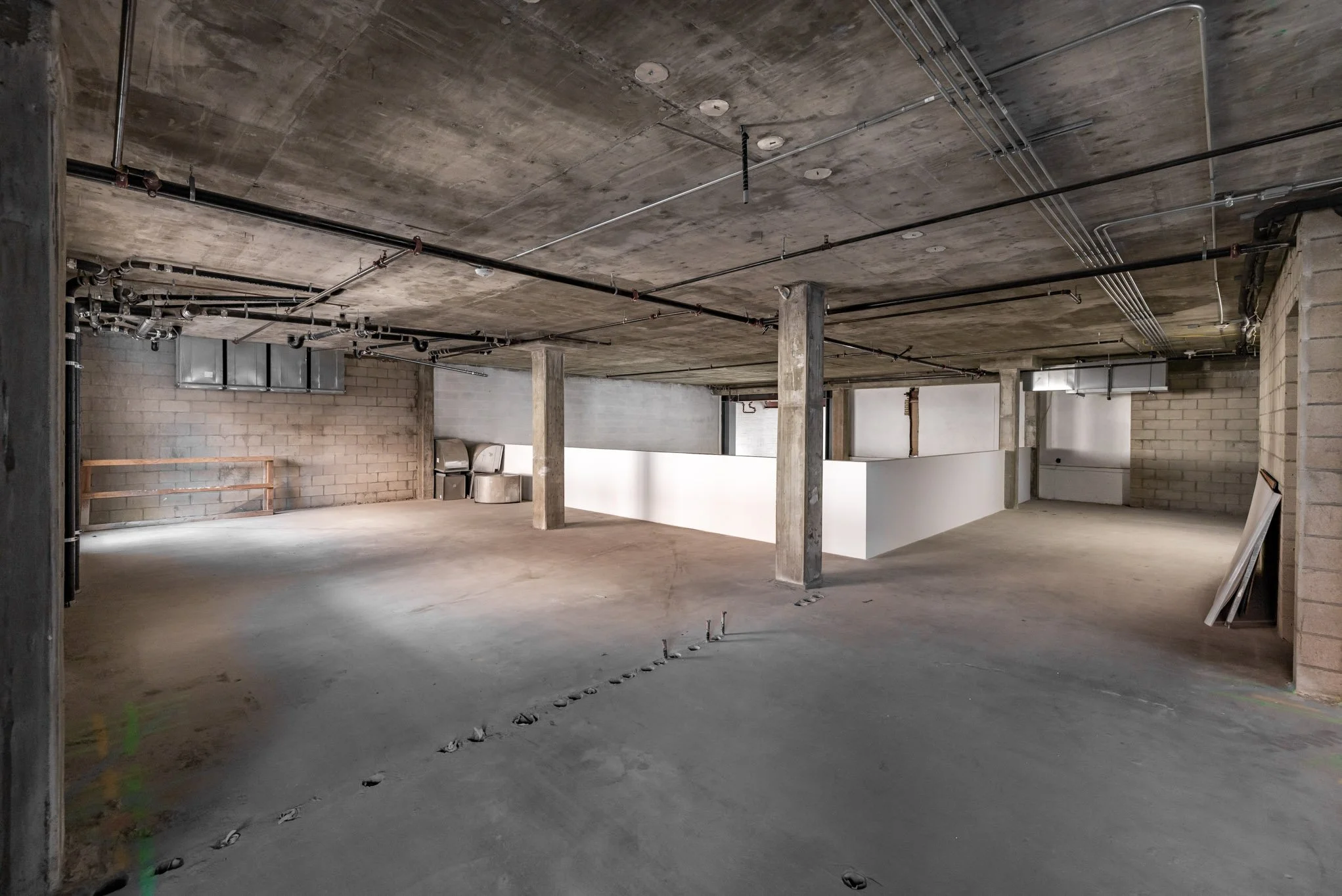 Interior view of an unfinished building with concrete floors, exposed ceiling pipes, and brick walls, showing open space and construction materials.