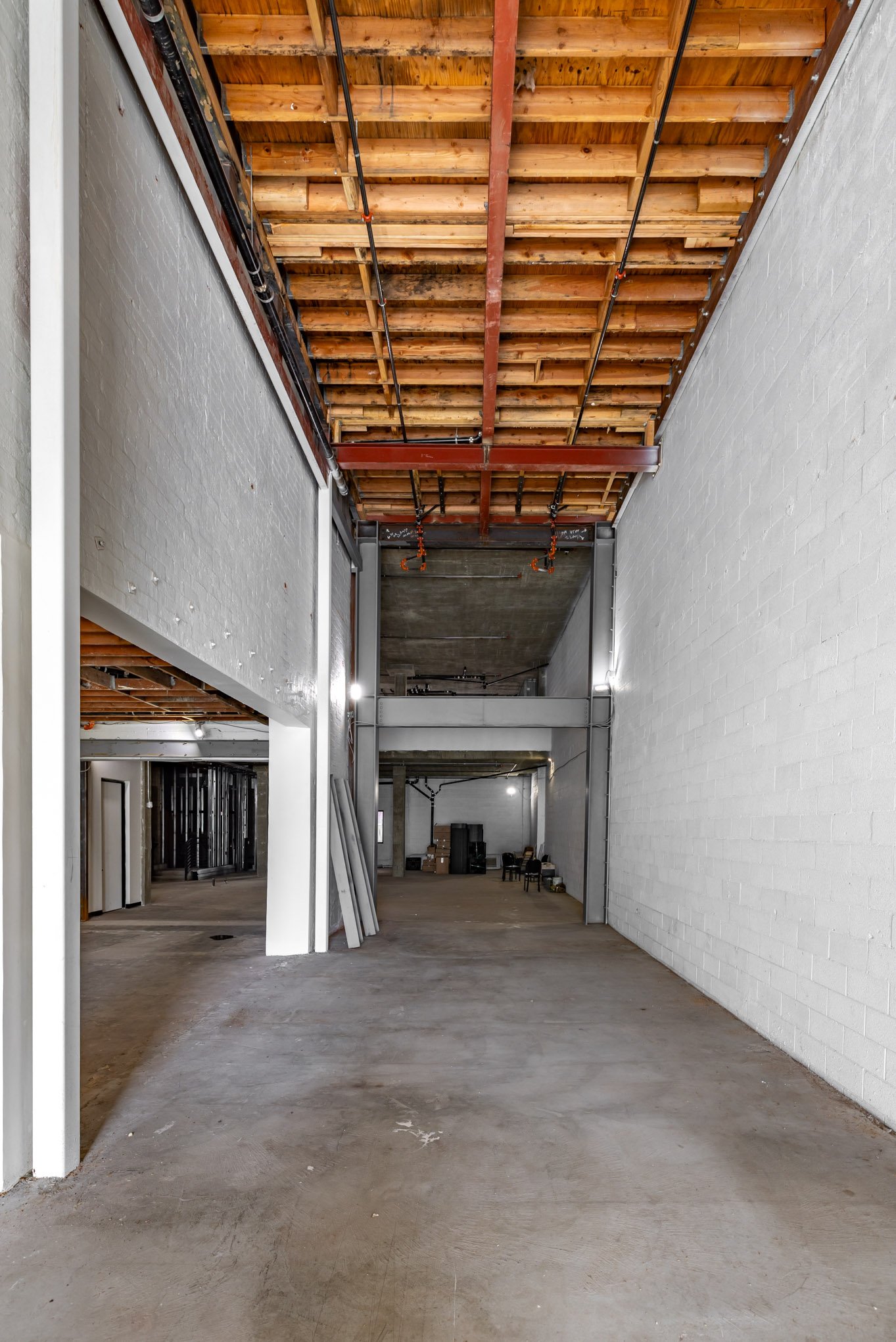 Interior view of a building under construction with an unfinished ceiling showing wooden beams, metal pipes, and some construction materials on the concrete floor.