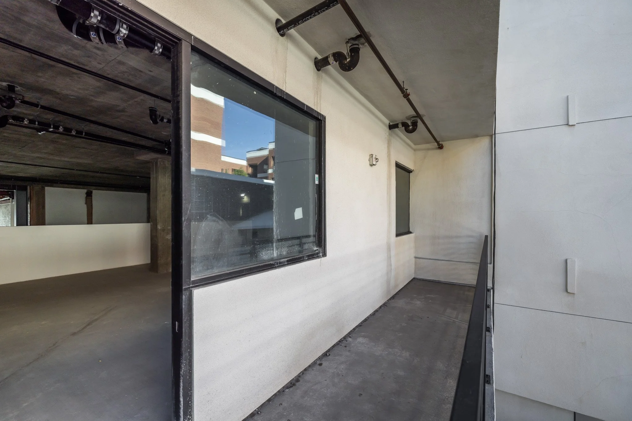 Empty apartment balcony with concrete flooring, white walls, and black metal railings, adjacent to windows and an unfinished interior.