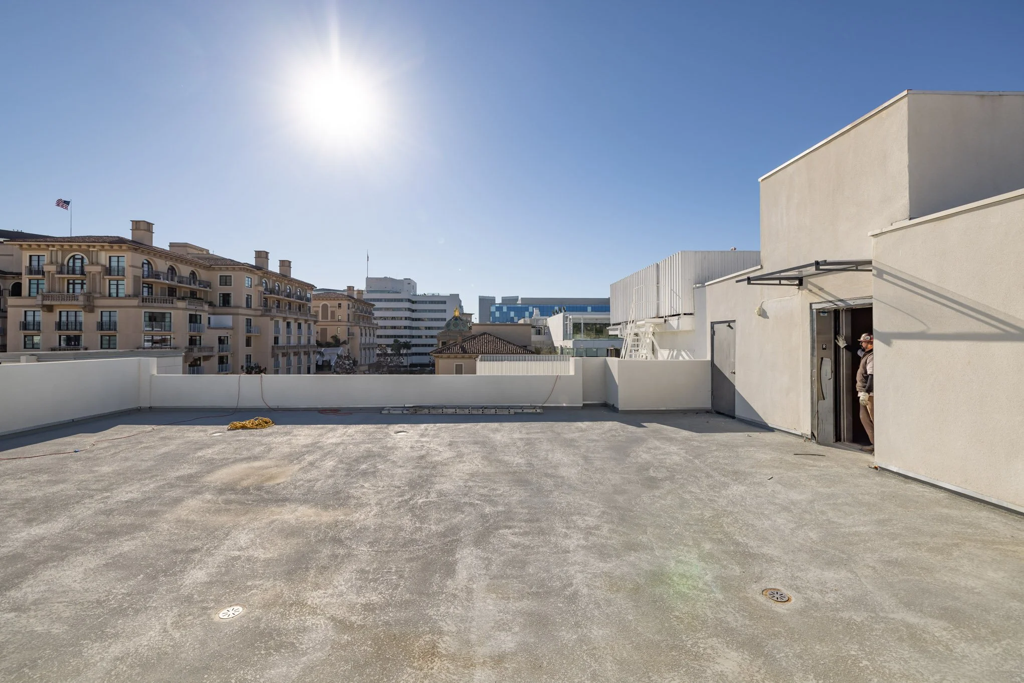 An empty rooftop with a person standing near the doorway on the right, overlooking a city skyline with multifamily residential buildings and office structures, under a bright sun in a clear blue sky.
