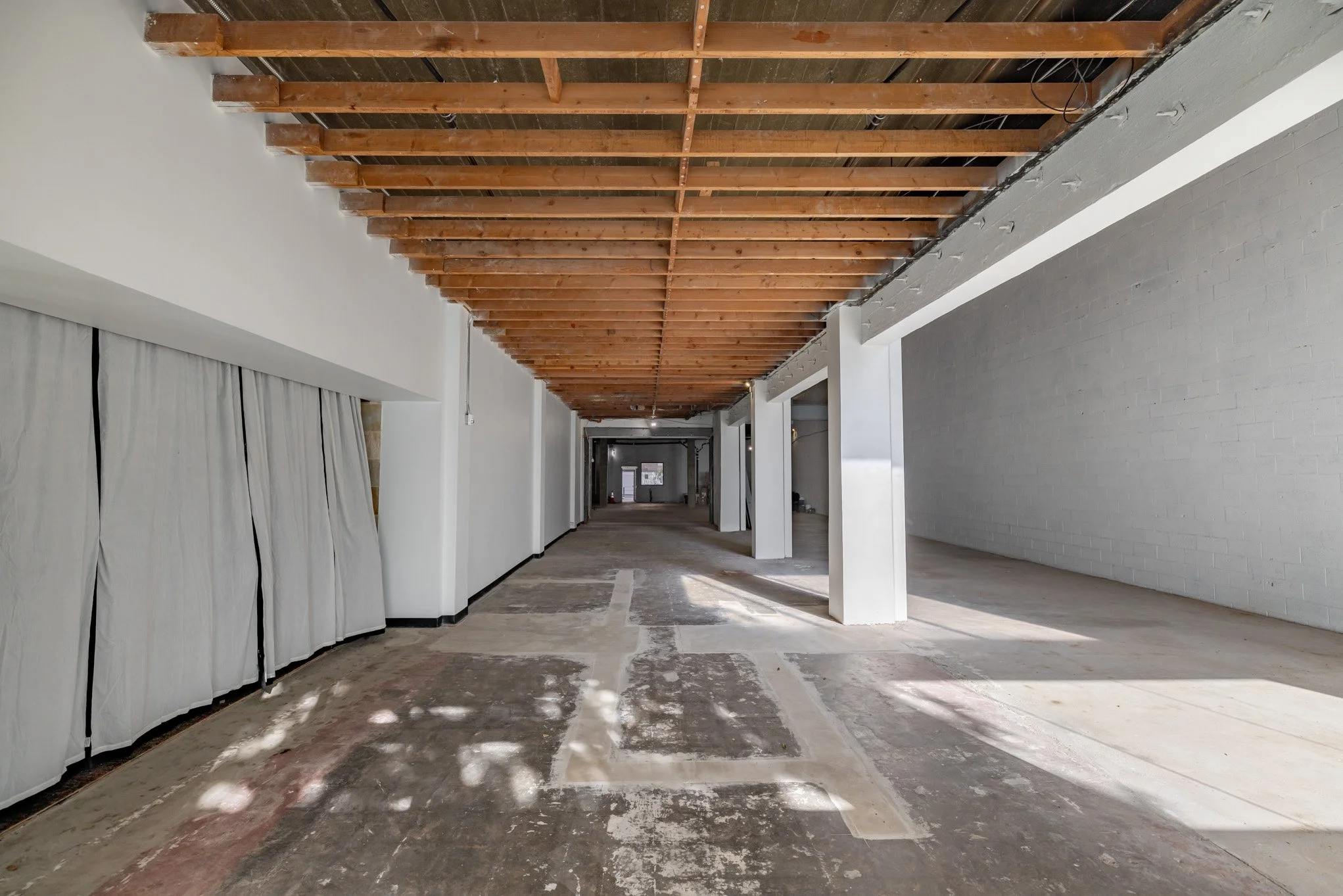 Empty interior space under construction with exposed wooden ceiling, white walls, and a concrete floor.