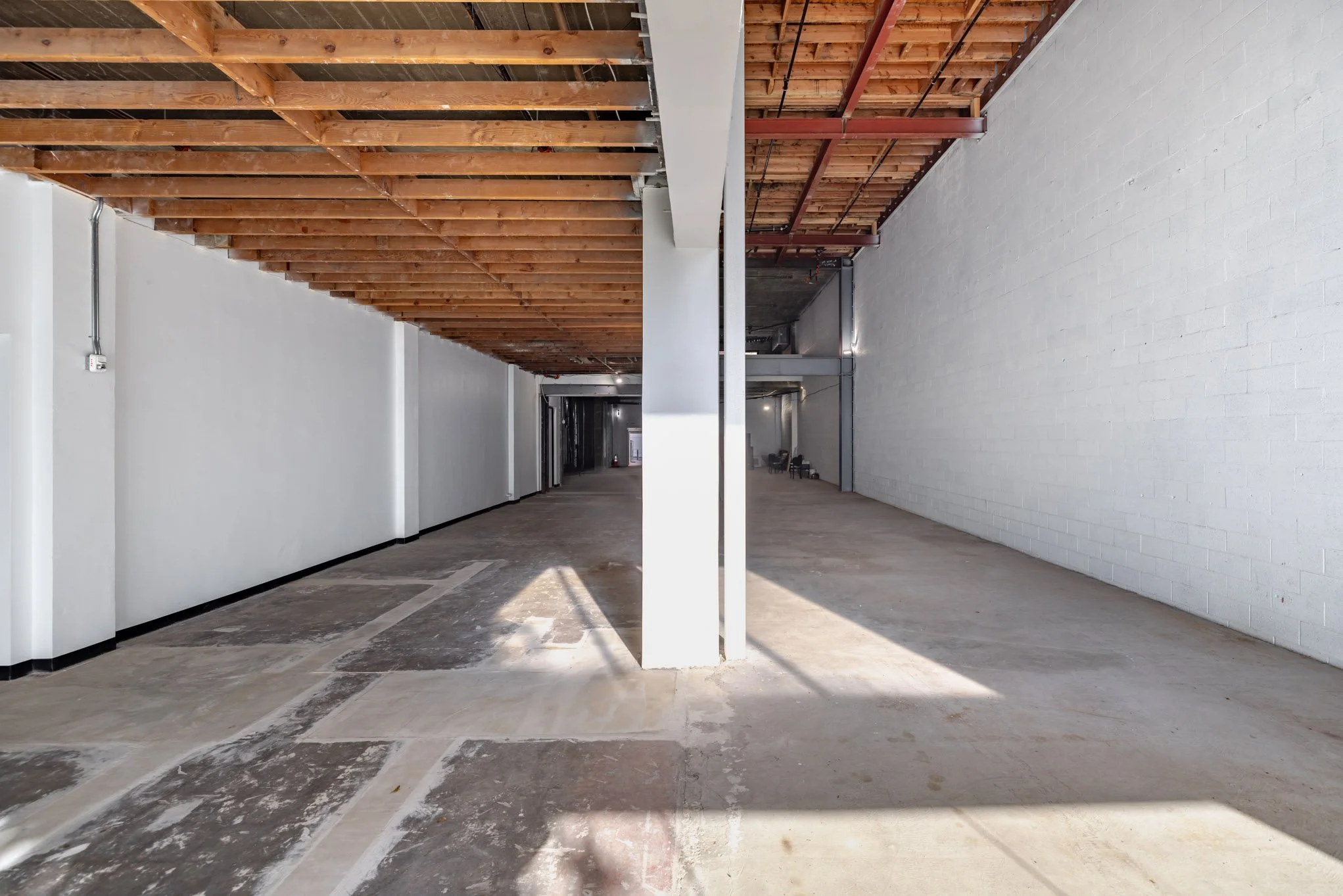 Empty interior of a commercial or industrial space under construction with exposed ceiling beams, white brick wall on the right, and a concrete floor.