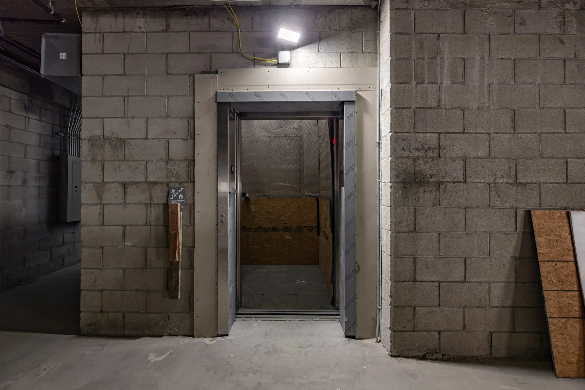 Construction site elevator door opening revealing plywood inside with unfinished concrete and brick walls around.
