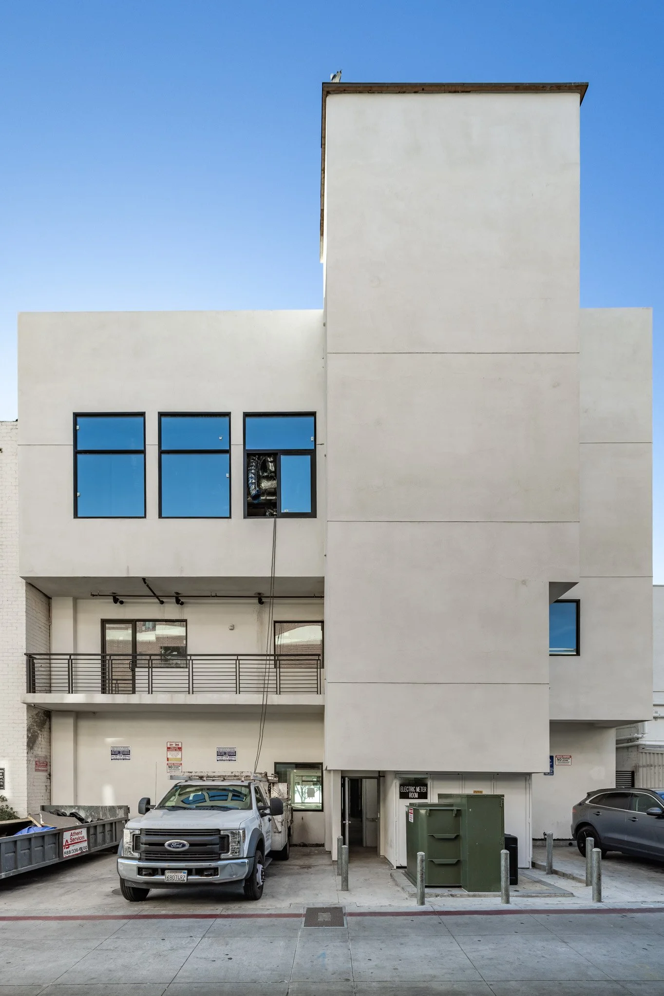 Modern white multi-story building with black window frames, a small balcony, parked vehicles, and a sign that says 'Electric Meter Room' in an urban setting.