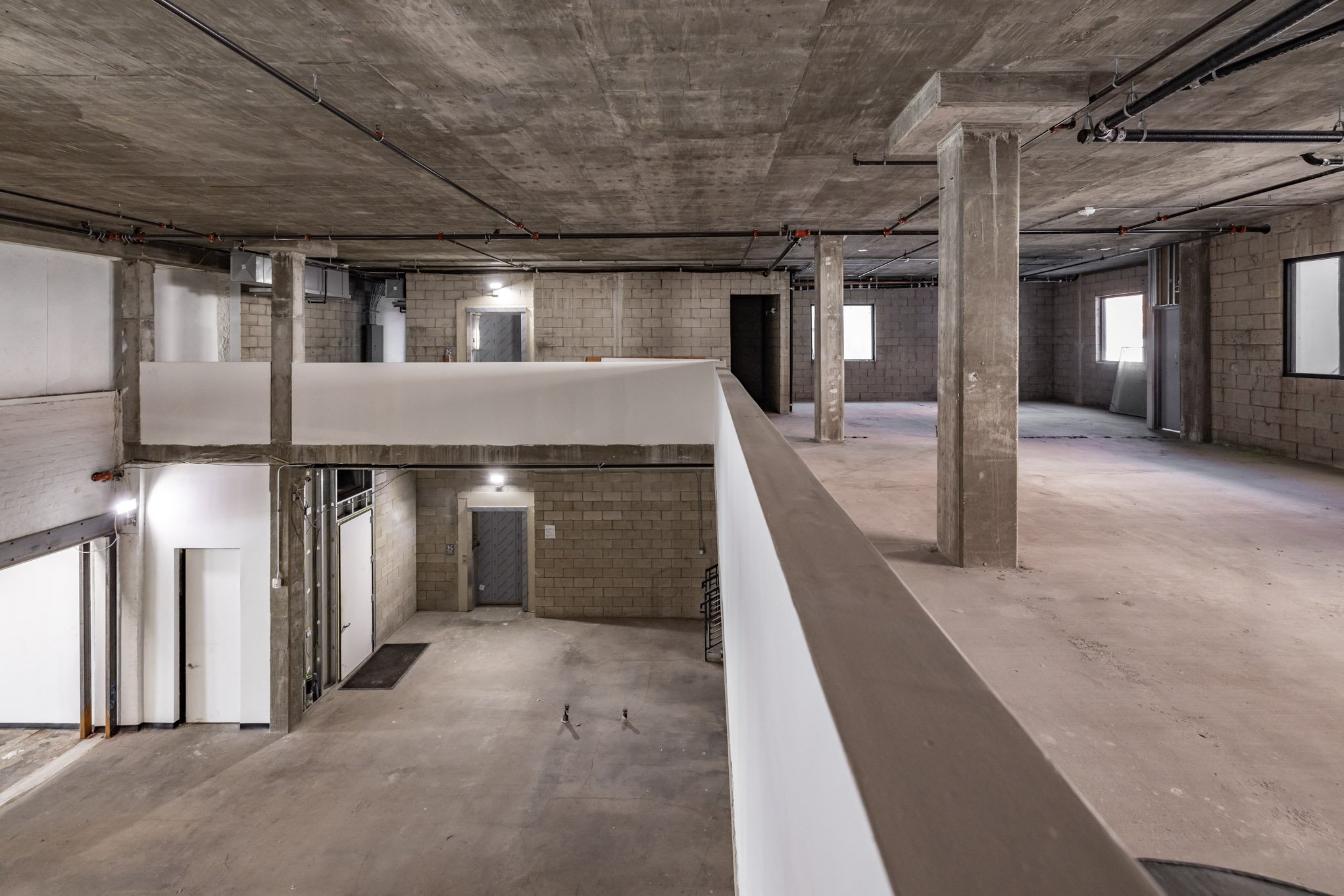 Interior of an unfinished construction building with exposed concrete columns, walls, and ceiling, and unfinished floors. Small windows are visible on the walls, and electrical pipes and wiring are installed across the ceiling.