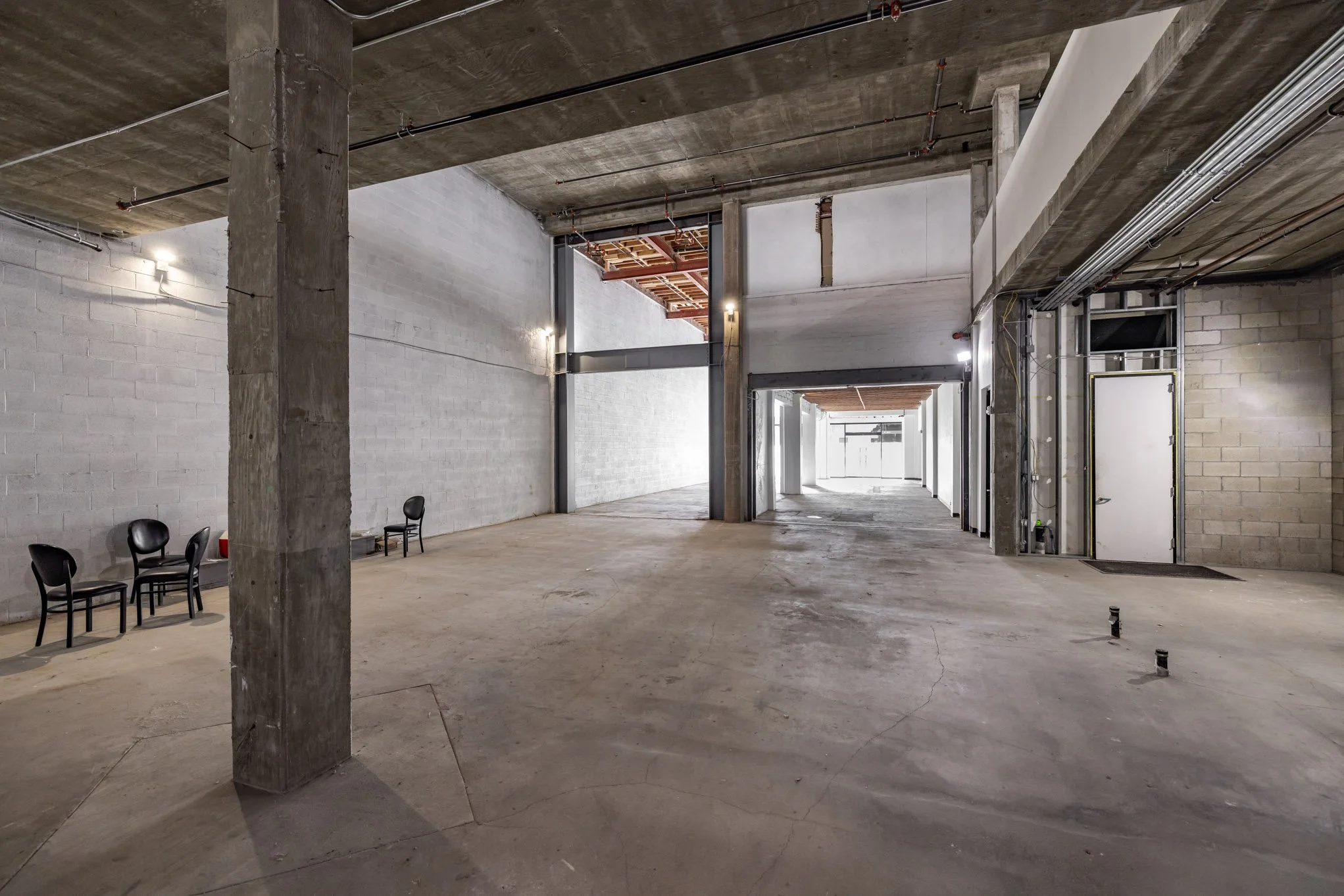 Interior view of an unfinished commercial or industrial space with concrete floors, exposed walls, and ceiling beams. There are three black chairs against the left wall and some construction materials and lighting on the right side.