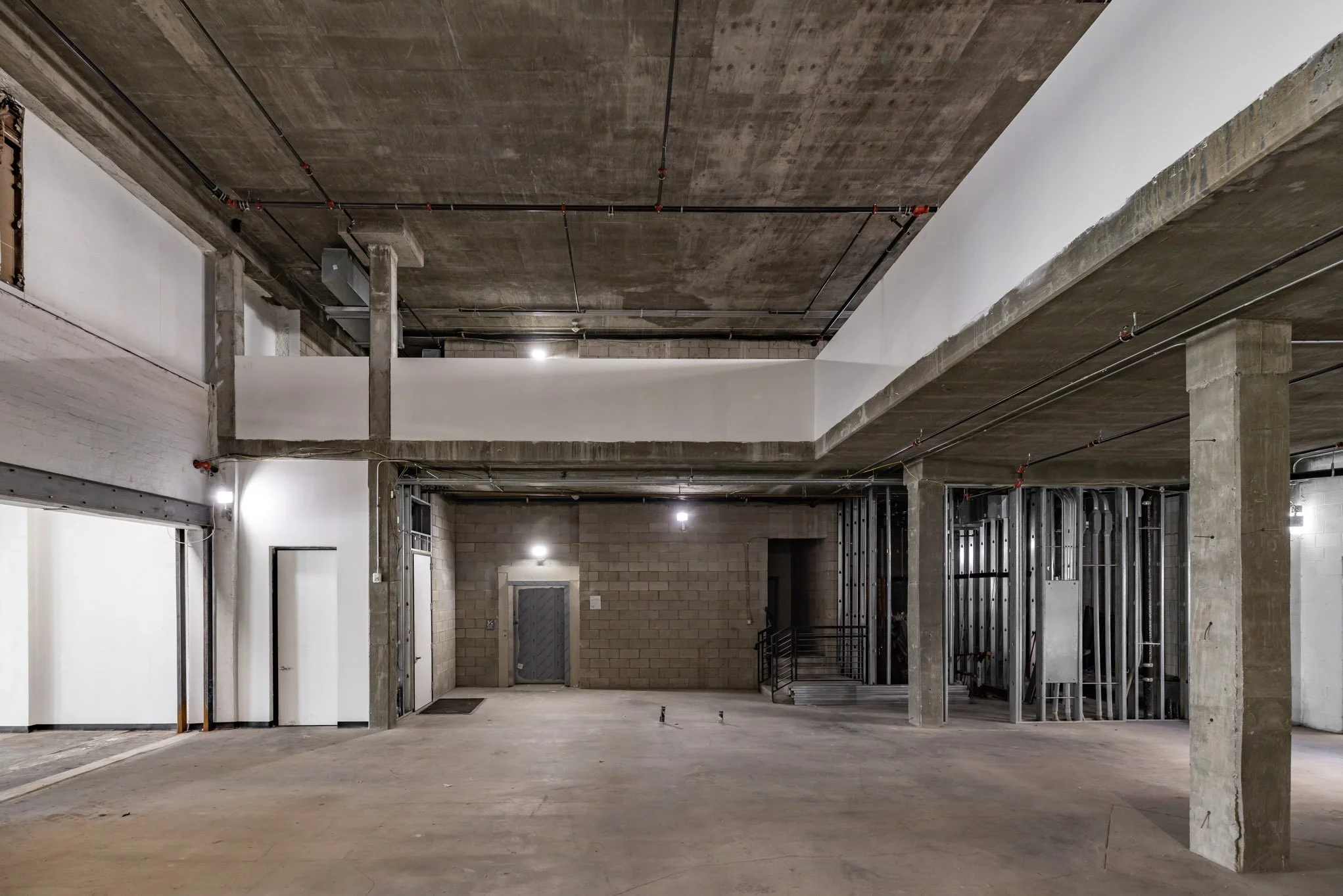 Empty, unfinished industrial space or parking garage with concrete floors, exposed concrete pillars, and partially finished walls. There are electrical conduits and lighting installed on the ceiling.