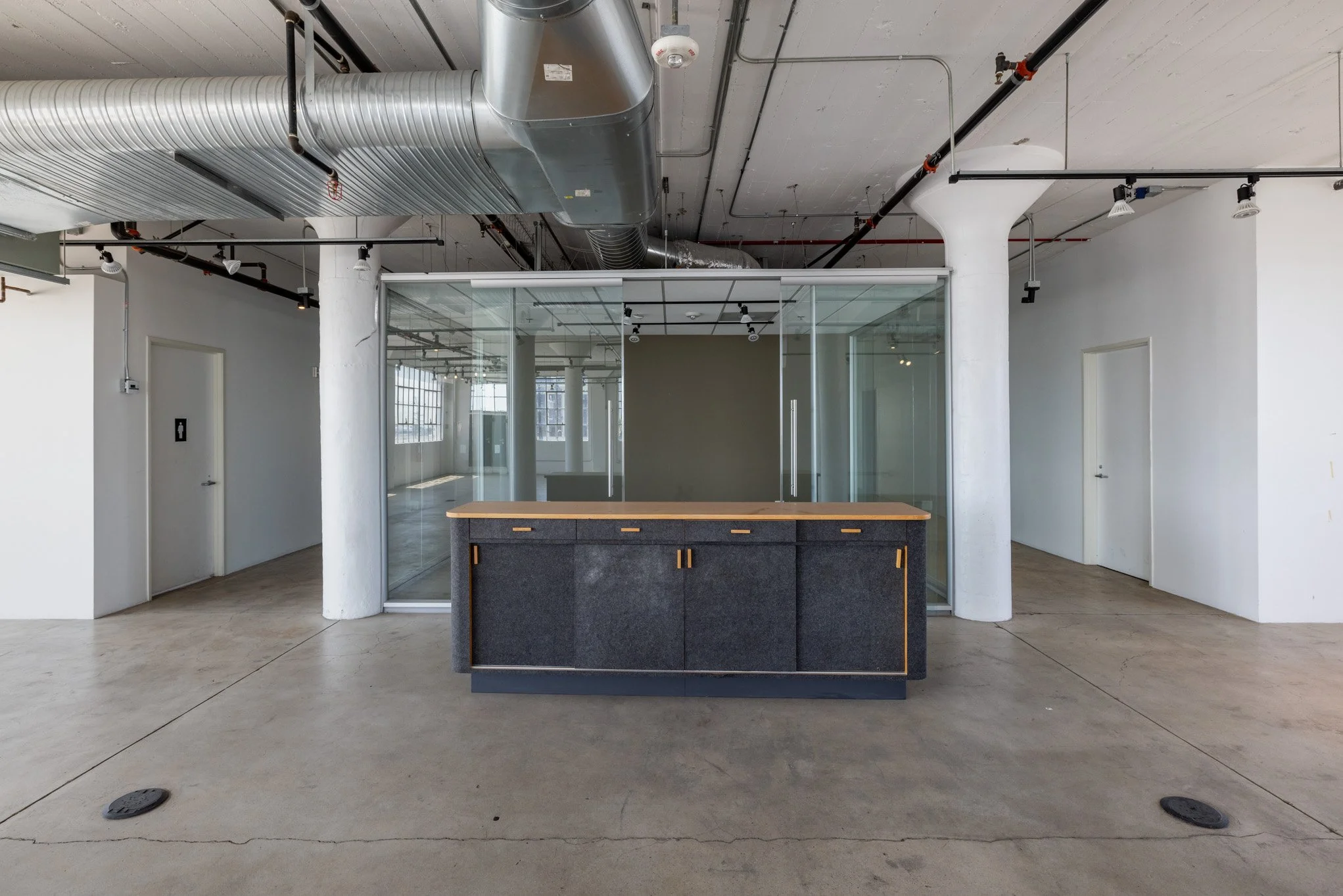 Interior view of a modern office space with a glass-walled conference room, an island with a dark base and wooden top, exposed ductwork on the ceiling, and white walls.