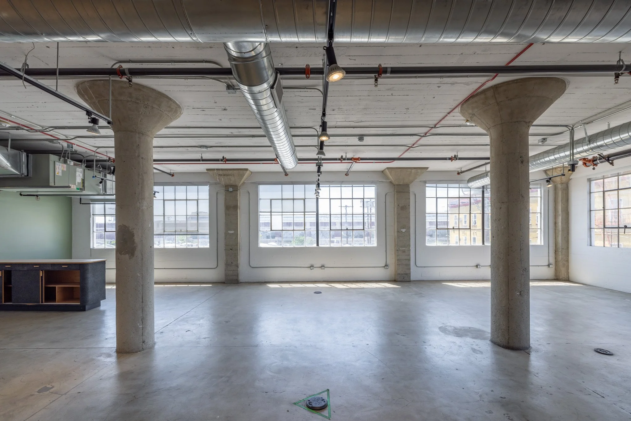 Empty industrial-style room with large windows, exposed concrete columns, and visible ductwork and pipes on the ceiling.