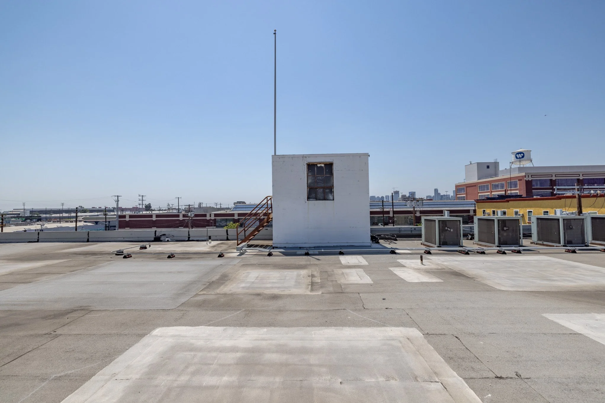 Rooftop view with a small white building, a staircase, multiple air conditioning units, and city buildings in the background under a clear blue sky.